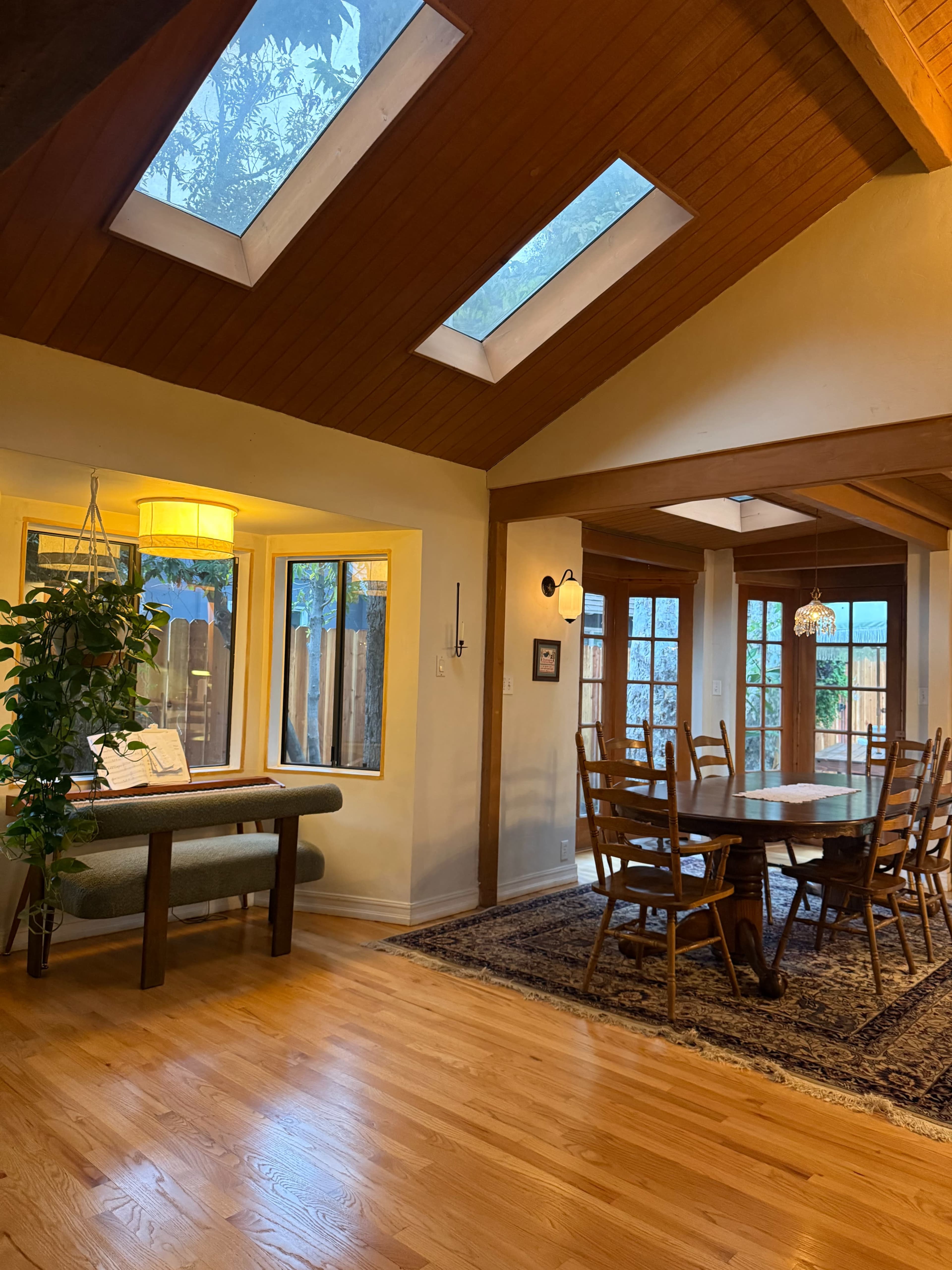 The image shows a well-lit dining area featuring a wooden table surrounded by chairs, with large windows and skylights allowing natural light to enter.