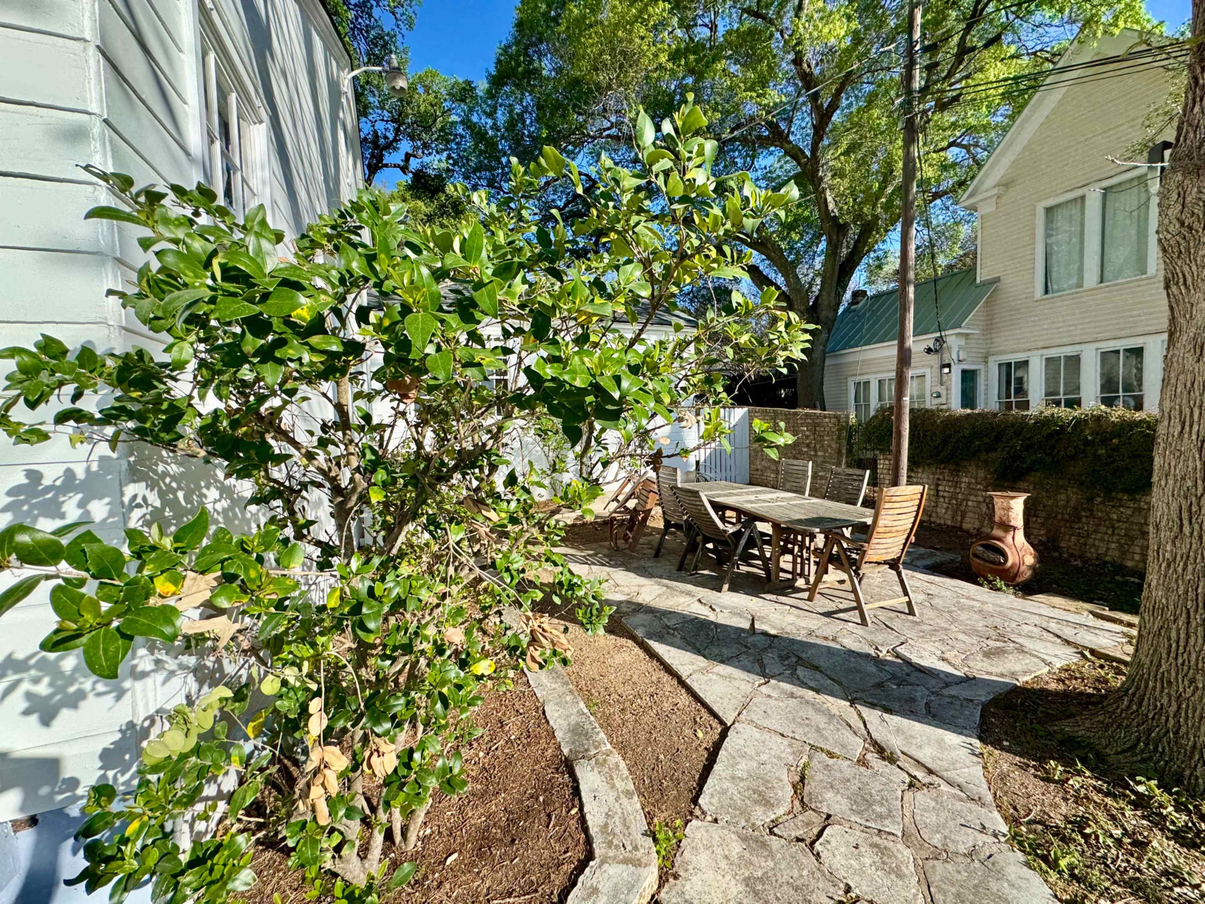 A stone patio with outdoor furniture is surrounded by greenery and residential houses in the background.