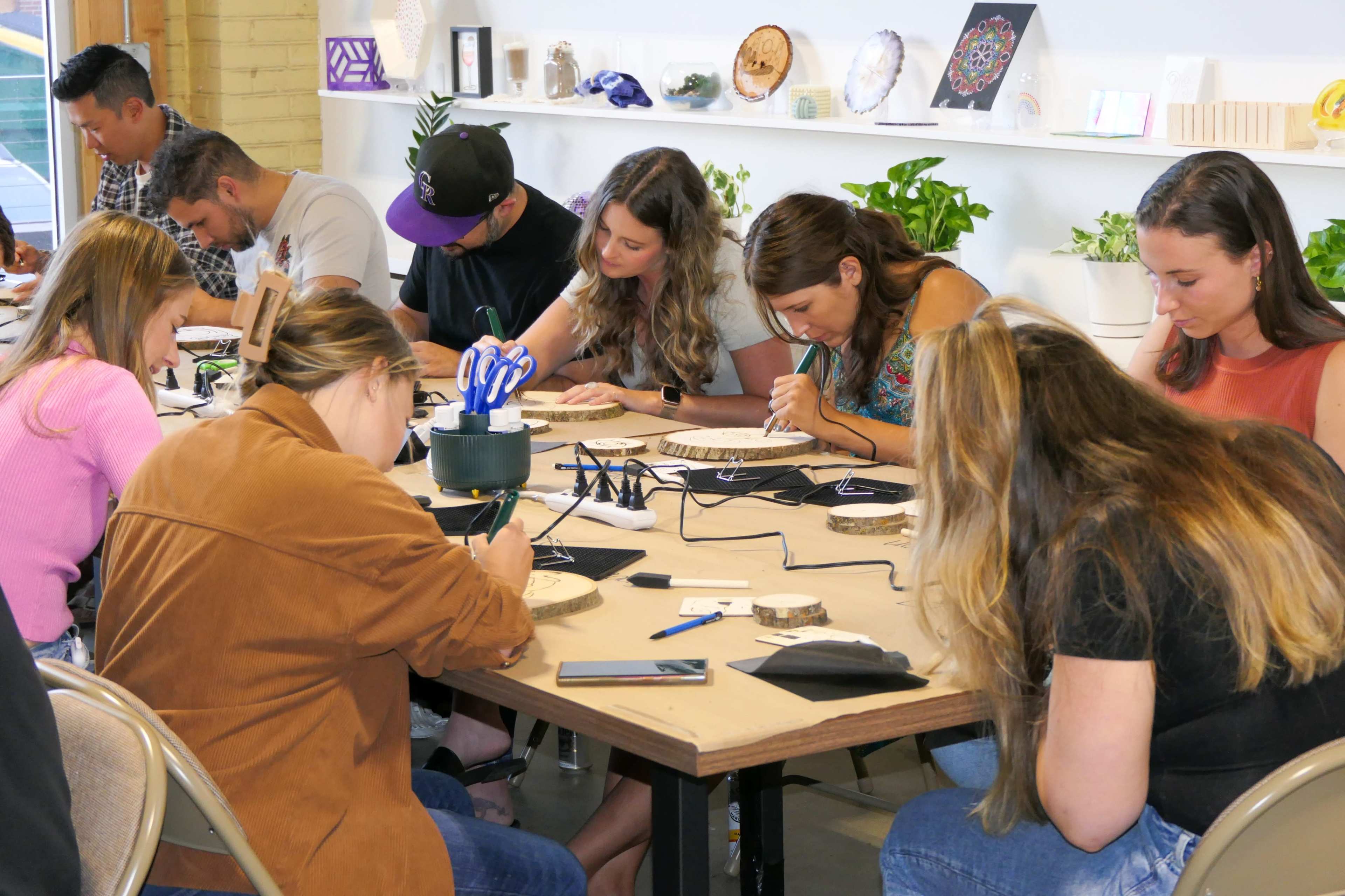 A group of eight people is focused on a crafting activity at a table, using tools to create designs on wooden pieces in a well-lit workspace.