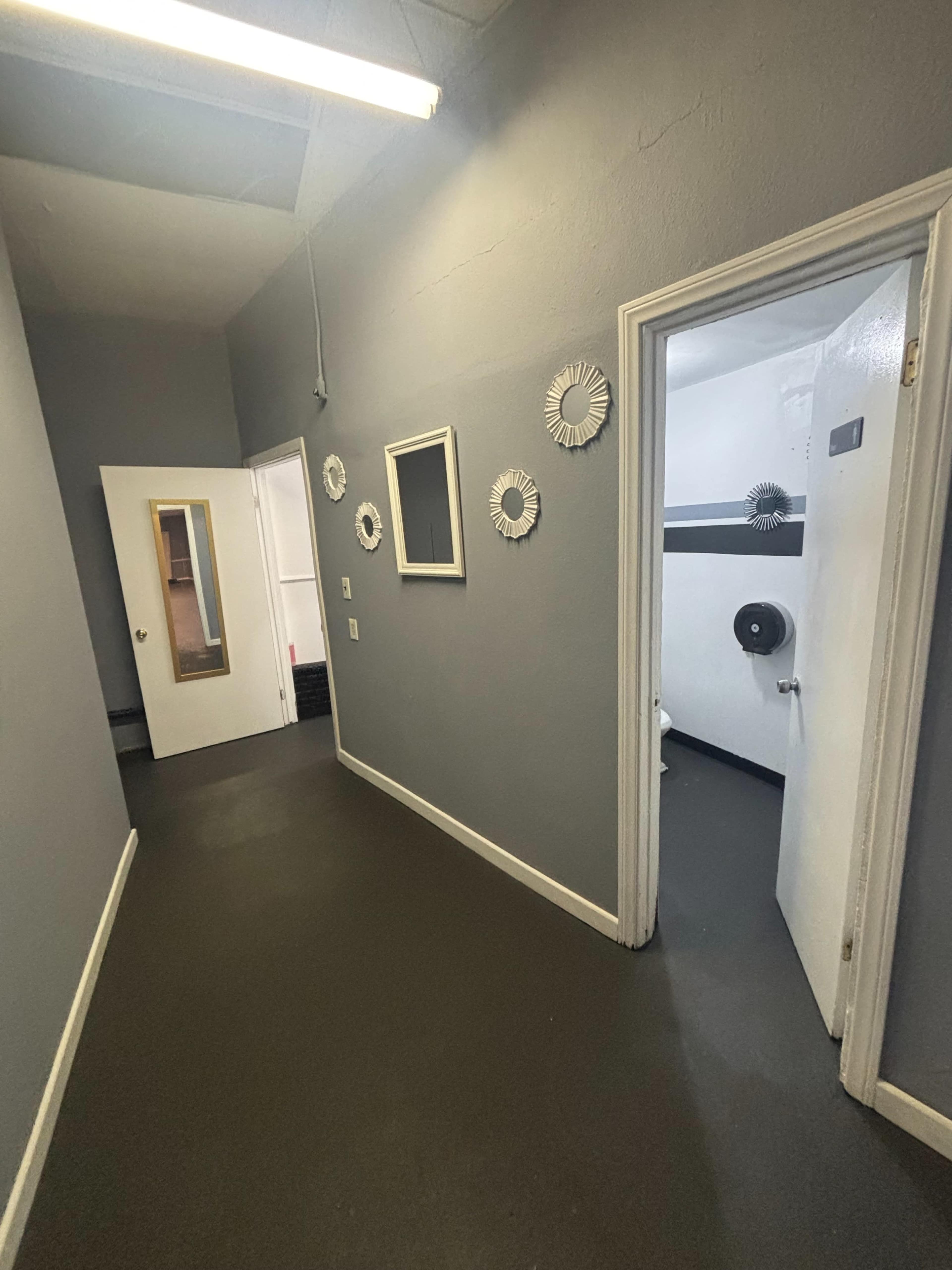 The image shows a dimly lit hallway with gray walls, a framed mirror on one side, and doors leading to other rooms at each end.