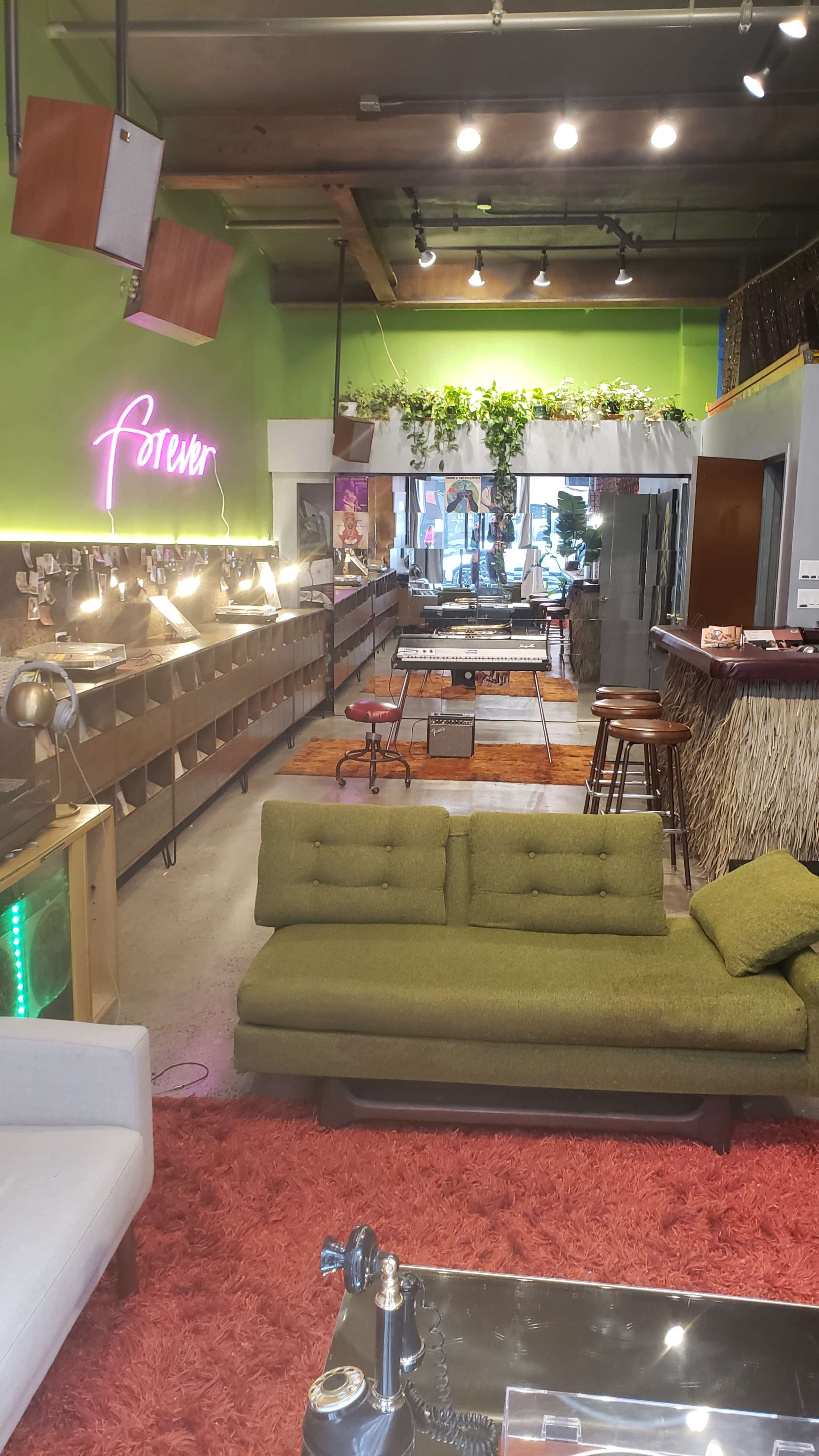 A cozy café interior featuring green walls, eclectic seating arrangements, and decorative plants suspended from the ceiling.
