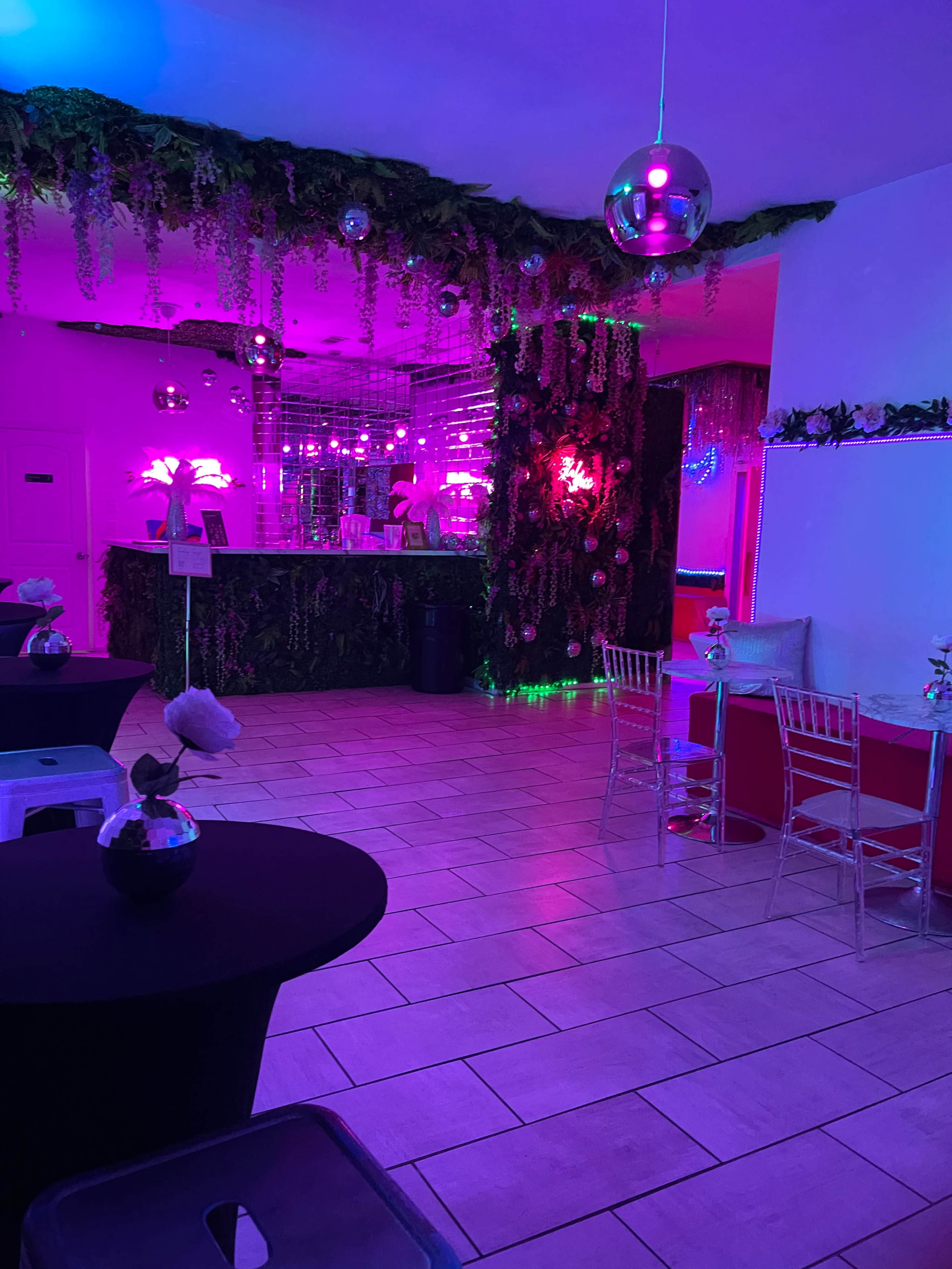 The image shows a brightly lit, colorful interior of a venue with plants and disco balls, featuring tables and a bar area.