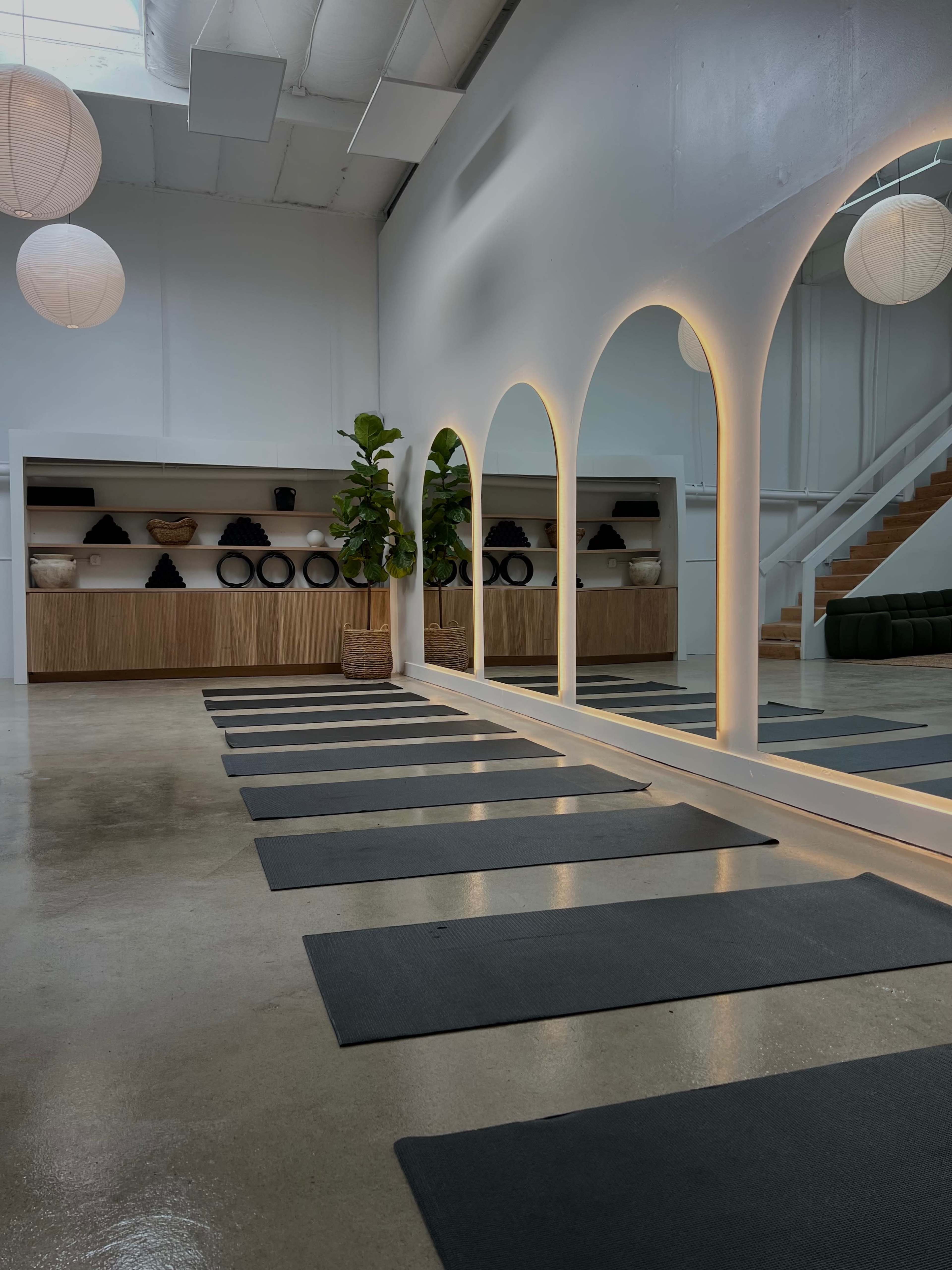 A spacious, minimalist studio with yoga mats arranged neatly on the floor, complemented by arch-shaped mirrors and a wooden shelf displaying decorative items.