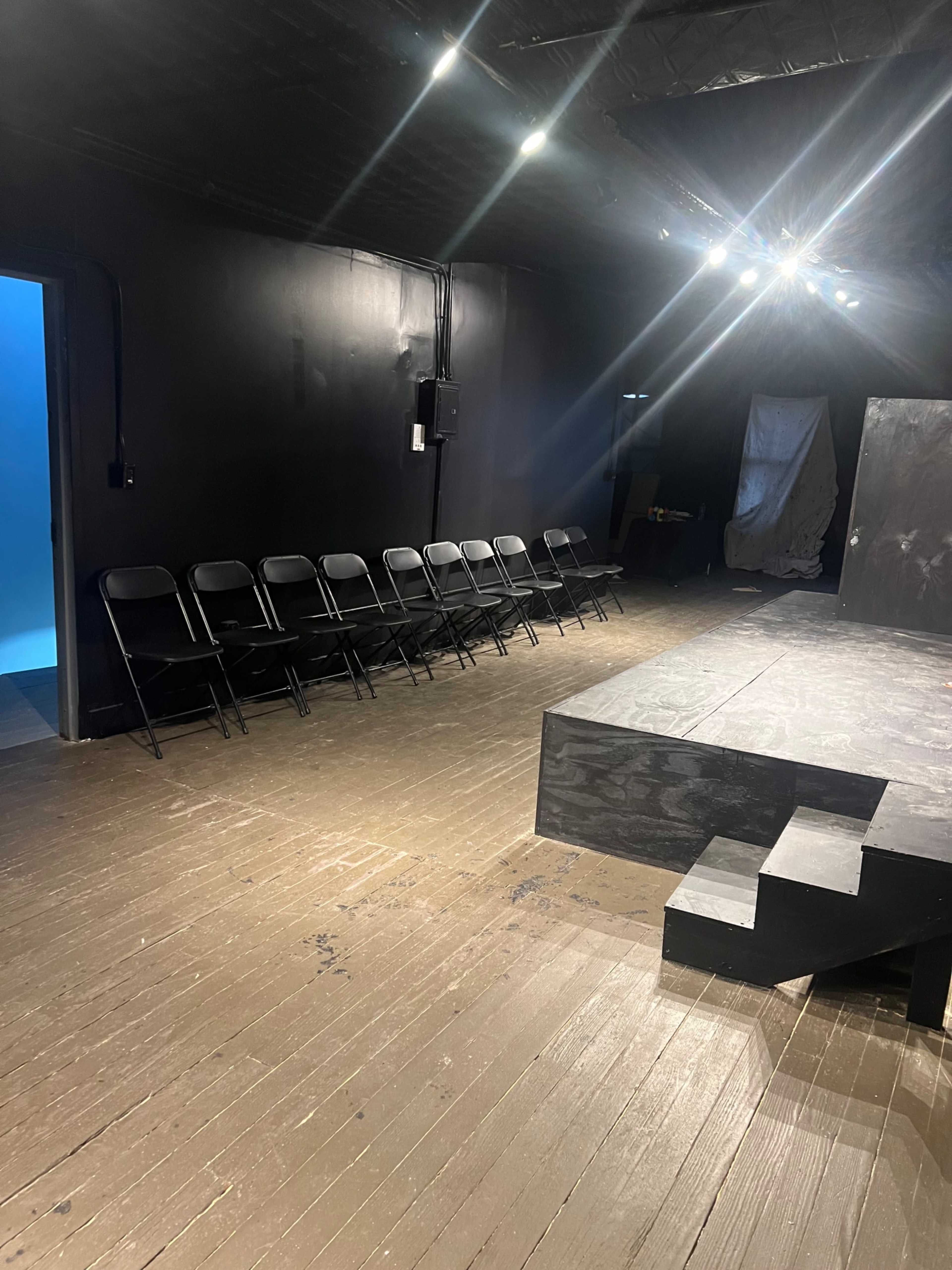 A black-walled theater space features a small stage with steps, several folding chairs arranged in a row, and a side door leading to another room.