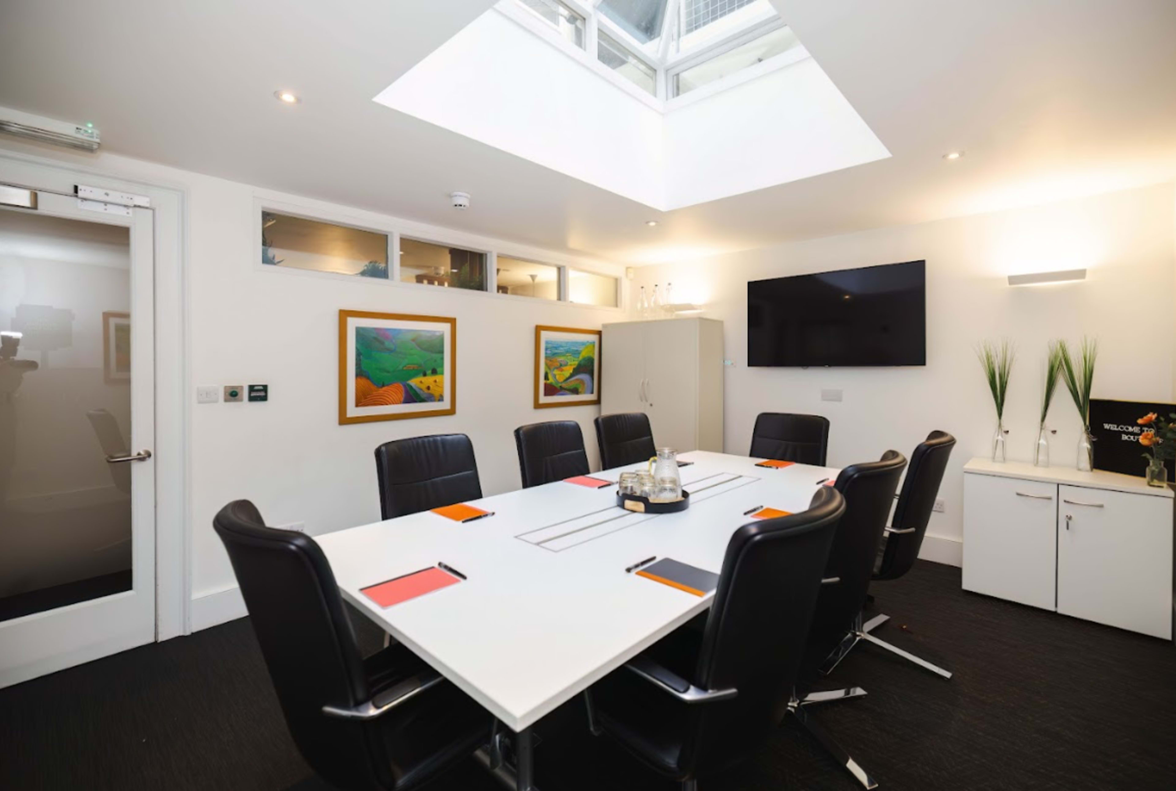 A modern conference room features a large white table surrounded by black chairs, with paintings on the walls and a skylight above.