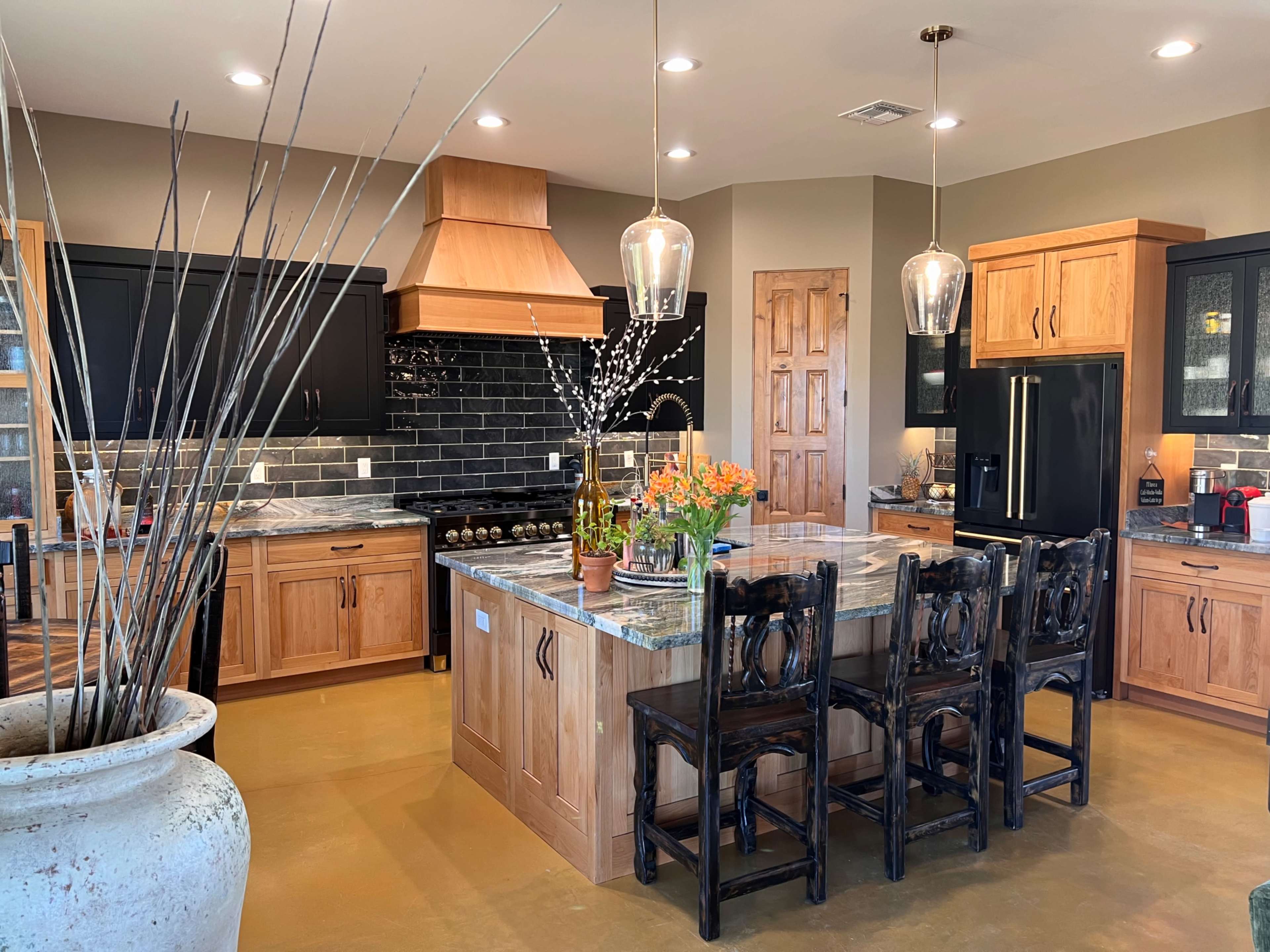 A modern kitchen with wooden cabinets, a large island with a stone countertop, and pendant lighting, featuring a mix of decorative plants and kitchen appliances.