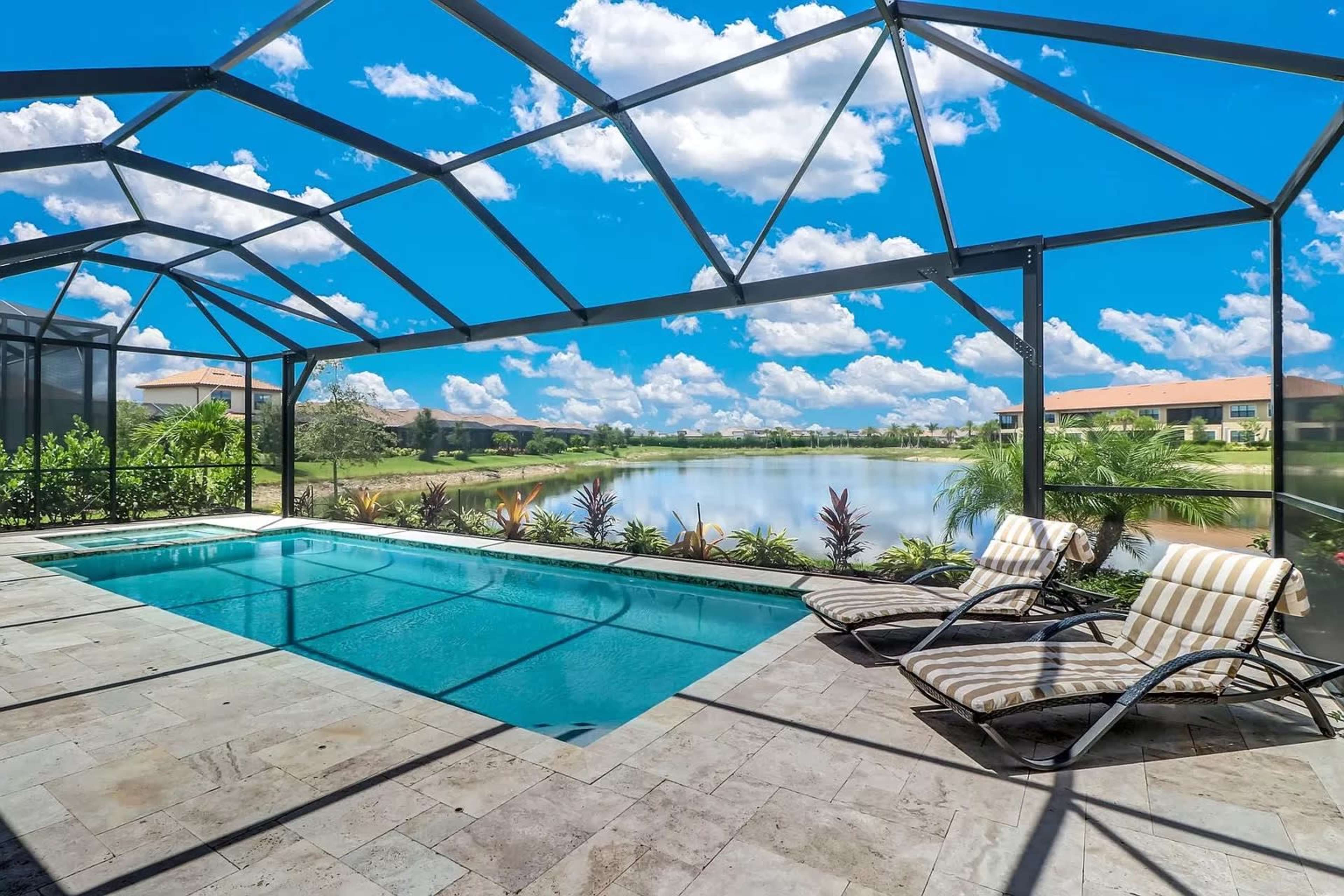 A screened pool area with two lounge chairs overlooks a calm lake surrounded by lush greenery under a bright sky.