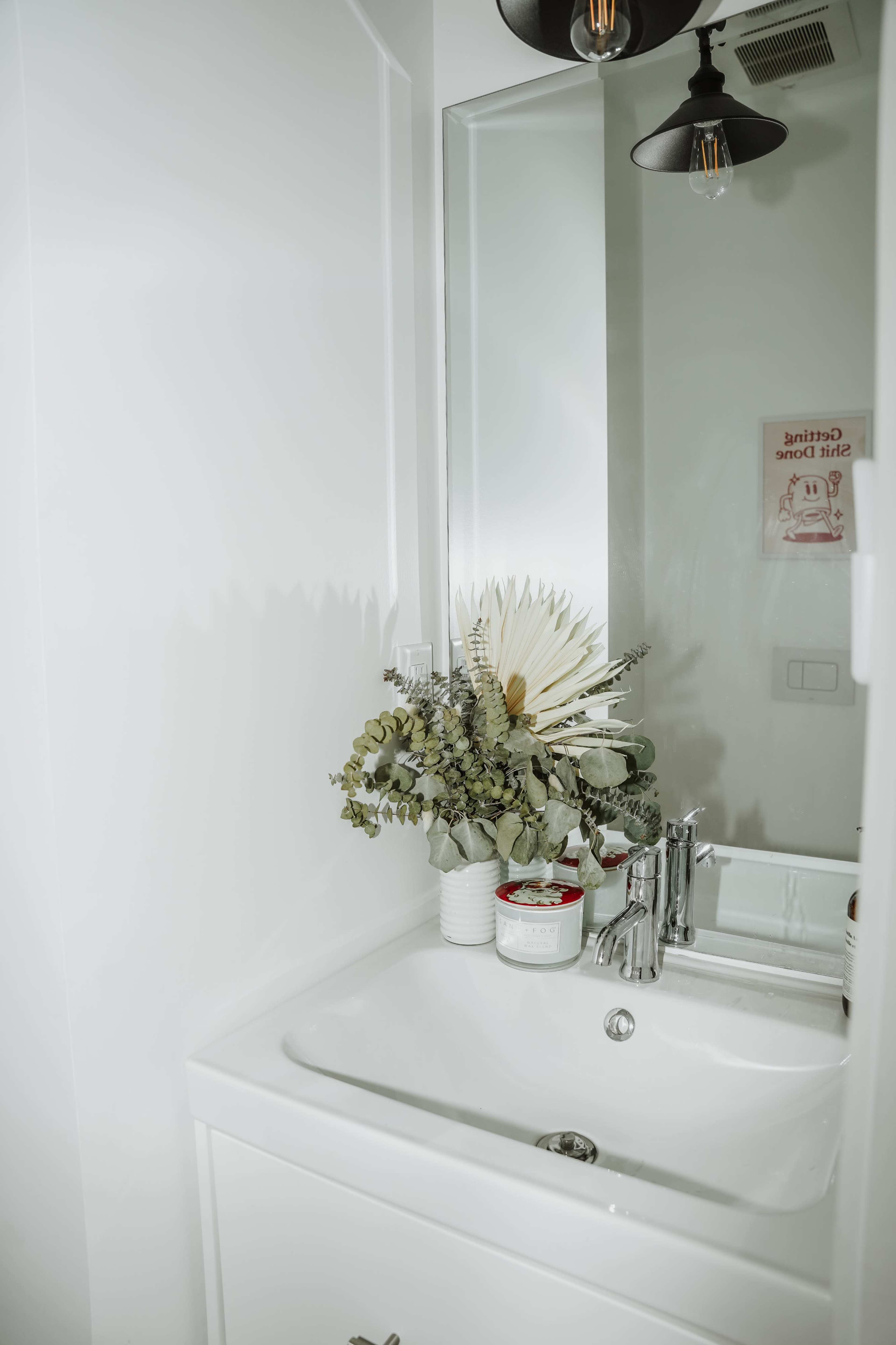 A modern bathroom features a white sink, a mirror reflecting a light fixture, and a decorative arrangement of dried plants on the countertop.