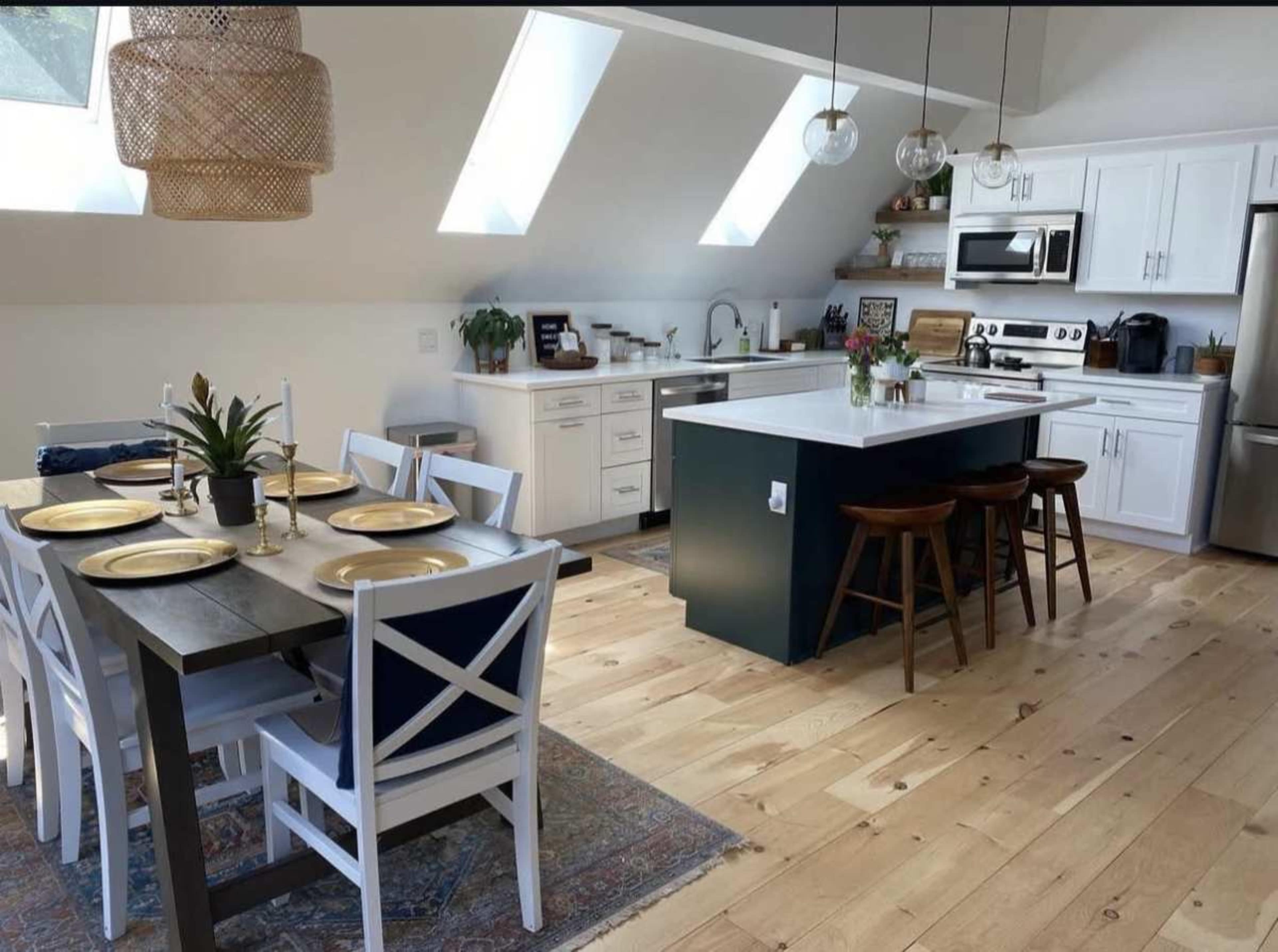 The image shows a modern kitchen and dining area with a table set for four, an island with bar stools, and skylights that allow natural light to flood the space.