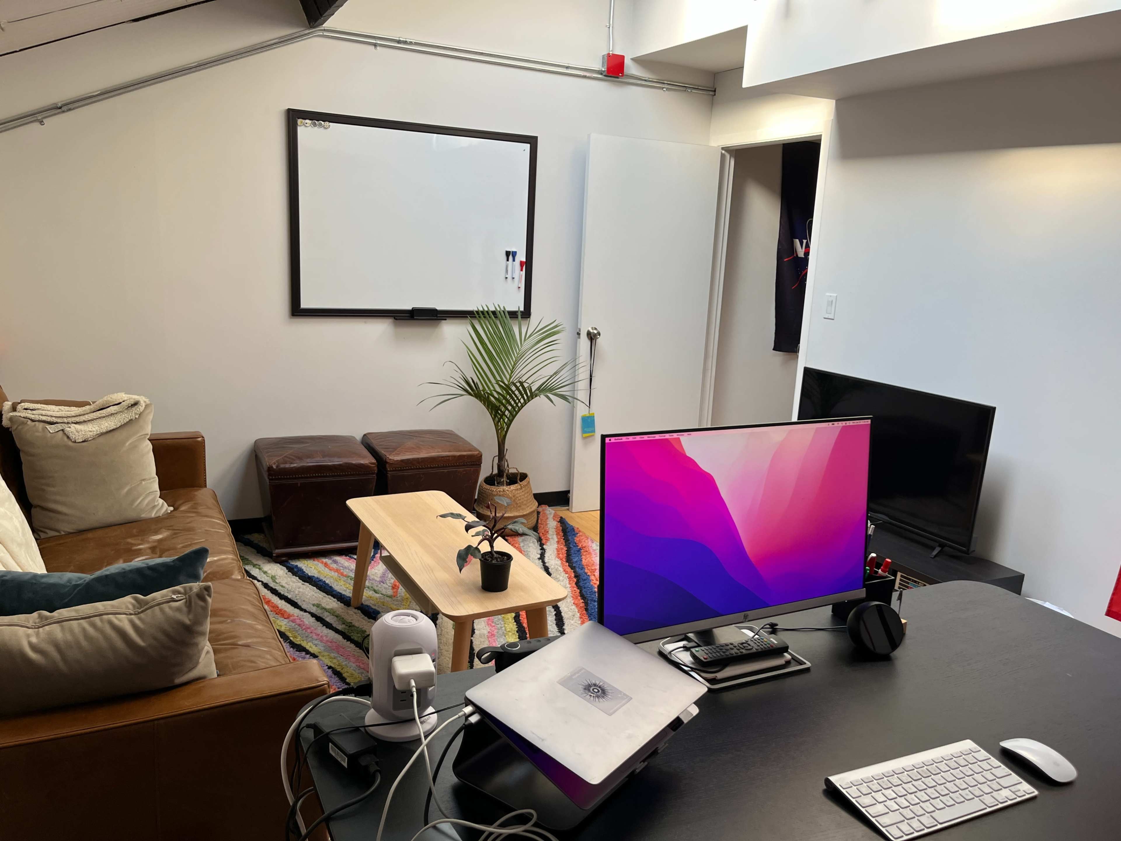 A modern home office space with a brown sofa, a wooden coffee table, a desktop computer, and a potted plant, all arranged on a colorful rug.