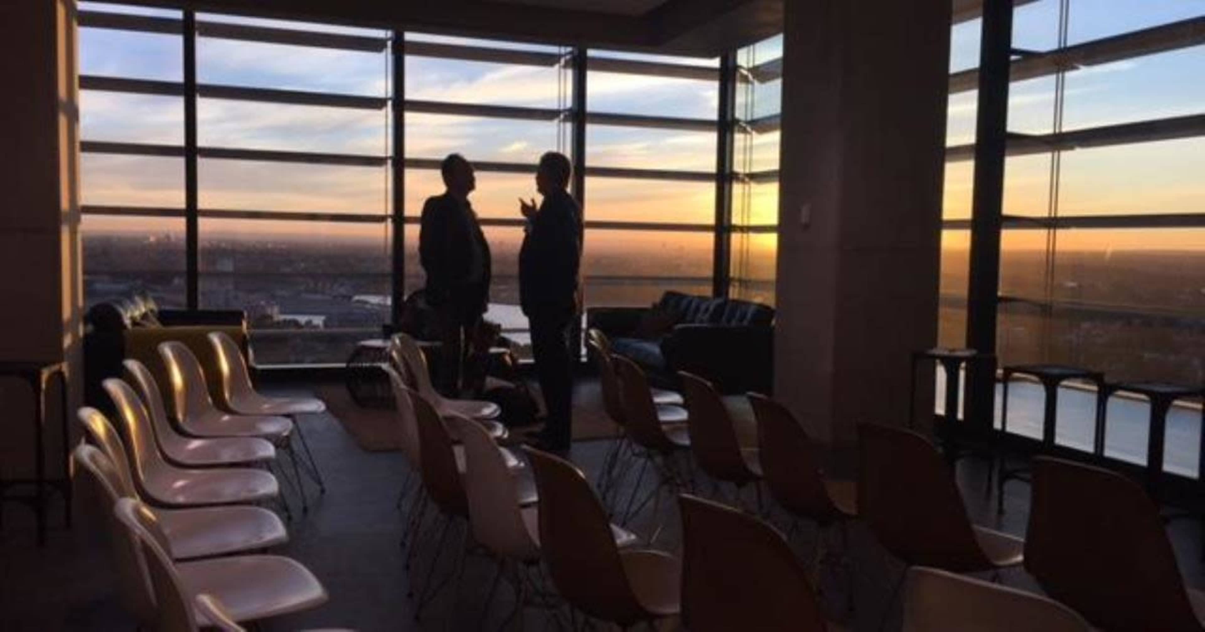 Two people stand and converse in a room with rows of empty chairs and large windows displaying a sunset view.