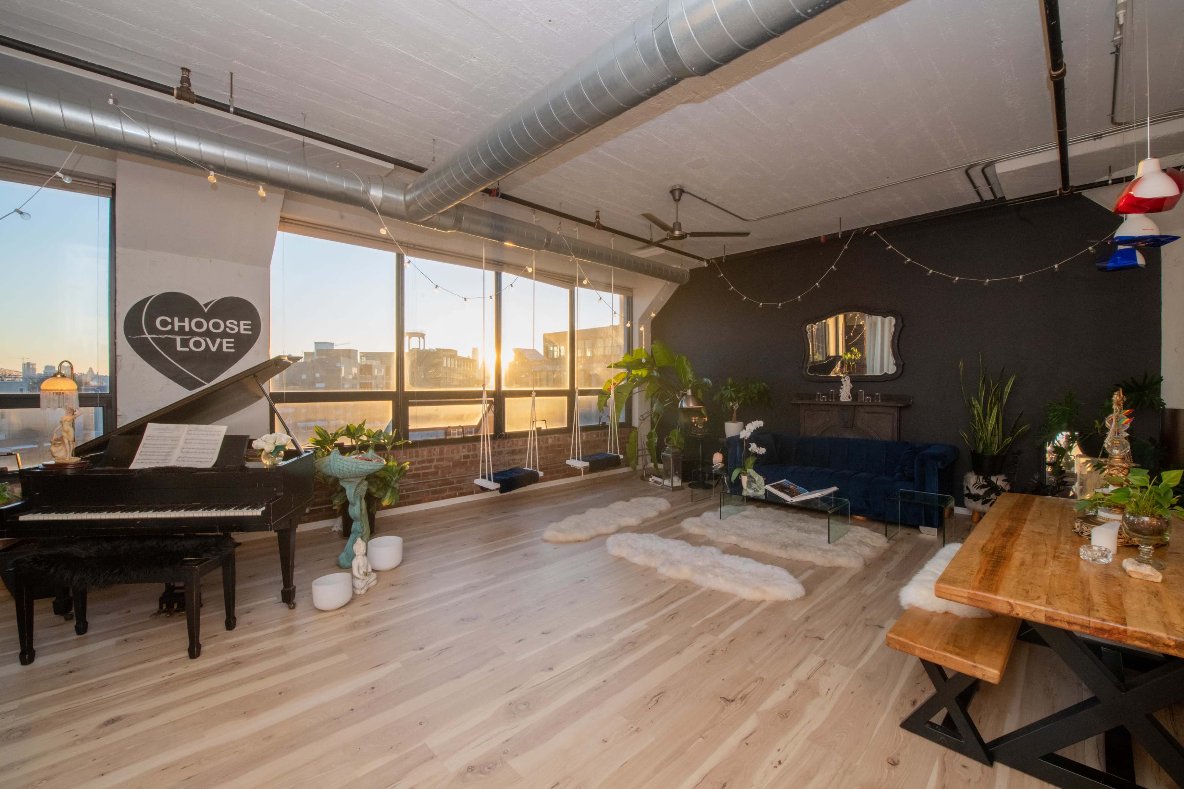 A spacious loft features a piano, a dark wall, a wooden dining table, and large windows showcasing a sunset view.