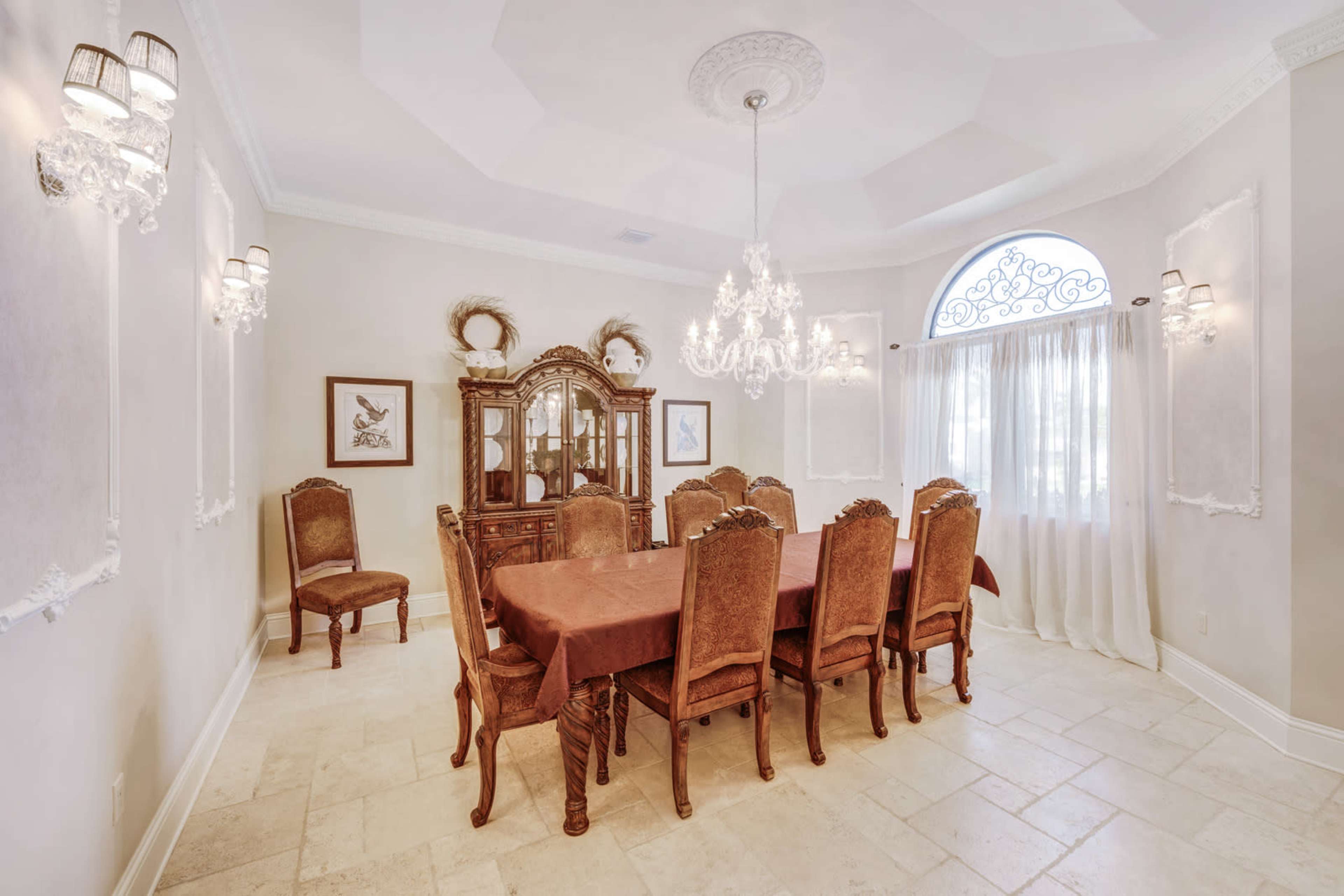 A dining room with a large wooden table surrounded by eight upholstered chairs, a chandelier hanging from the ceiling, and an ornate sideboard against the wall.