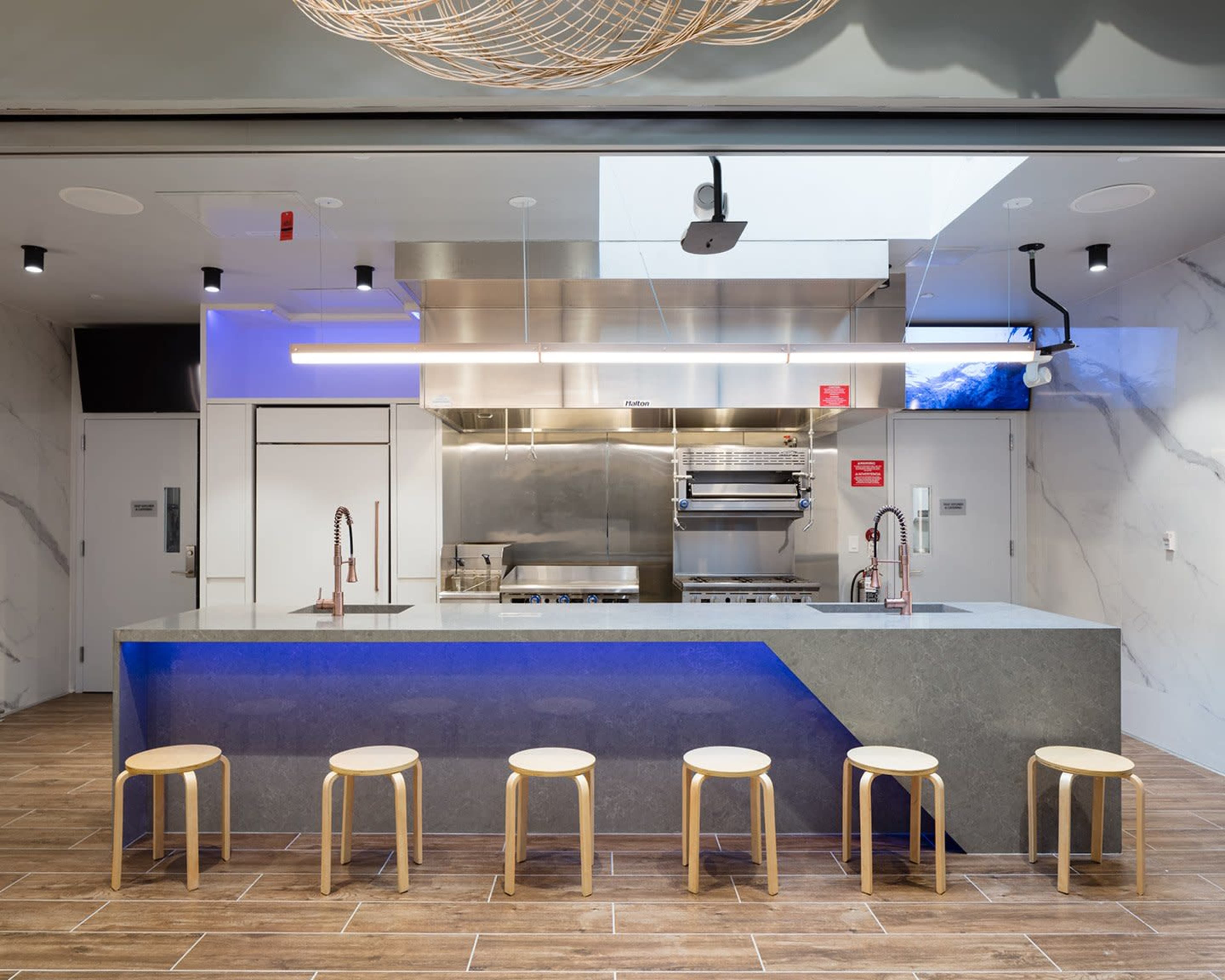 A modern kitchen with a large island and six wooden stools, featuring stainless steel appliances and a blue-lit countertop.