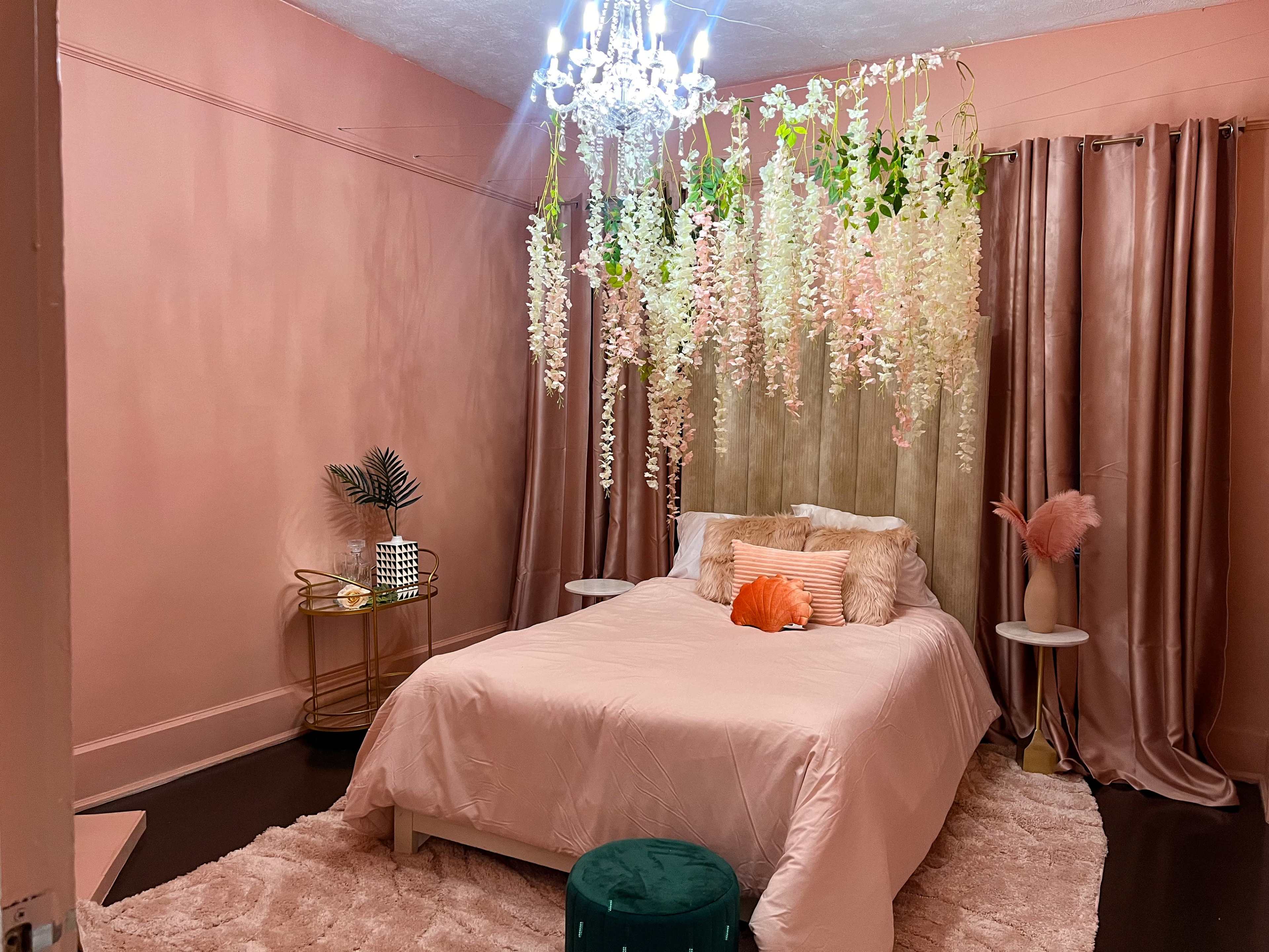 The image shows a room with a bed covered in a light pink comforter, surrounded by hanging floral decorations and curtains, with a plush rug and decorative side tables.