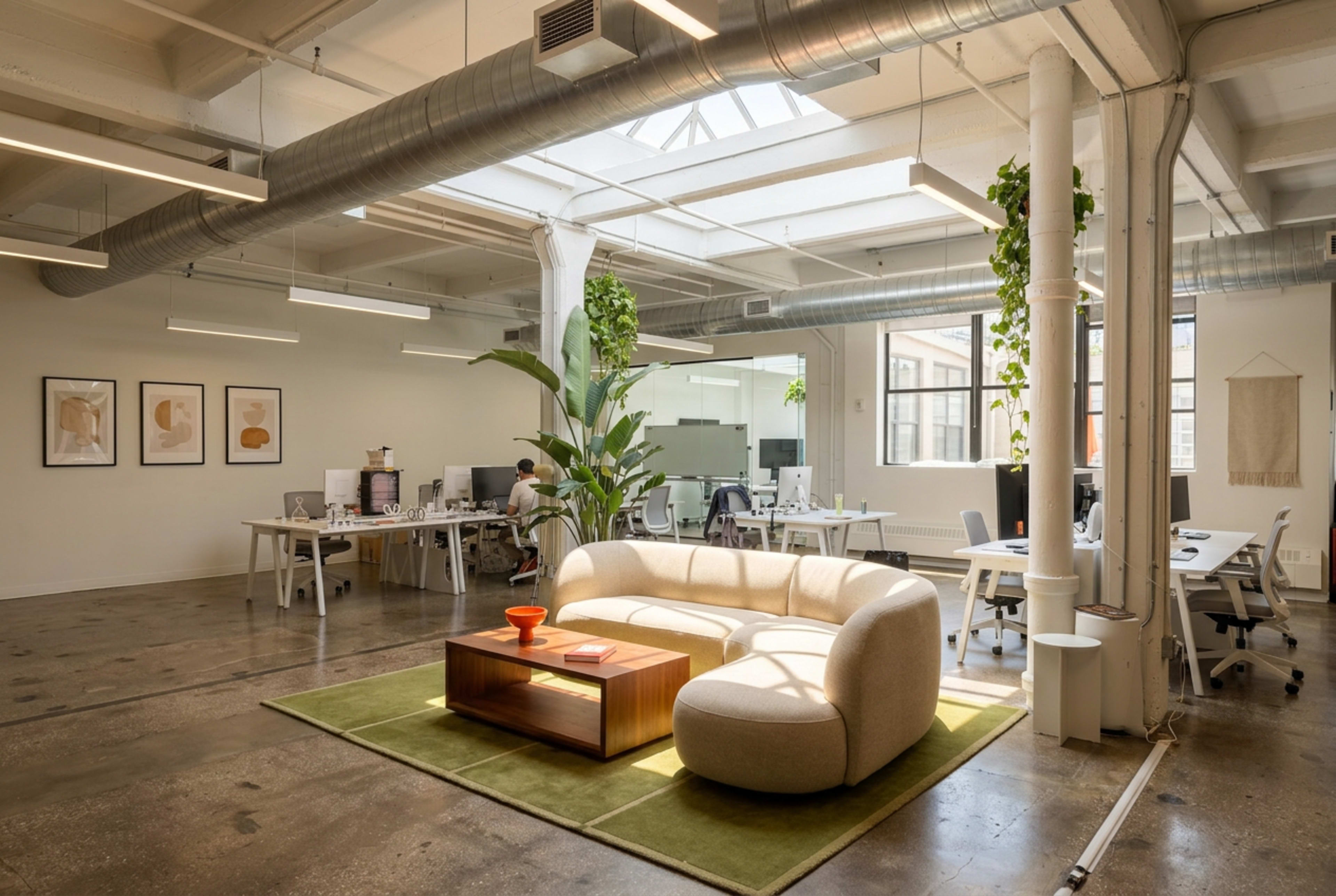 A spacious modern office features a curved sofa on a green rug, surrounded by desks and plants, with large windows letting in natural light.