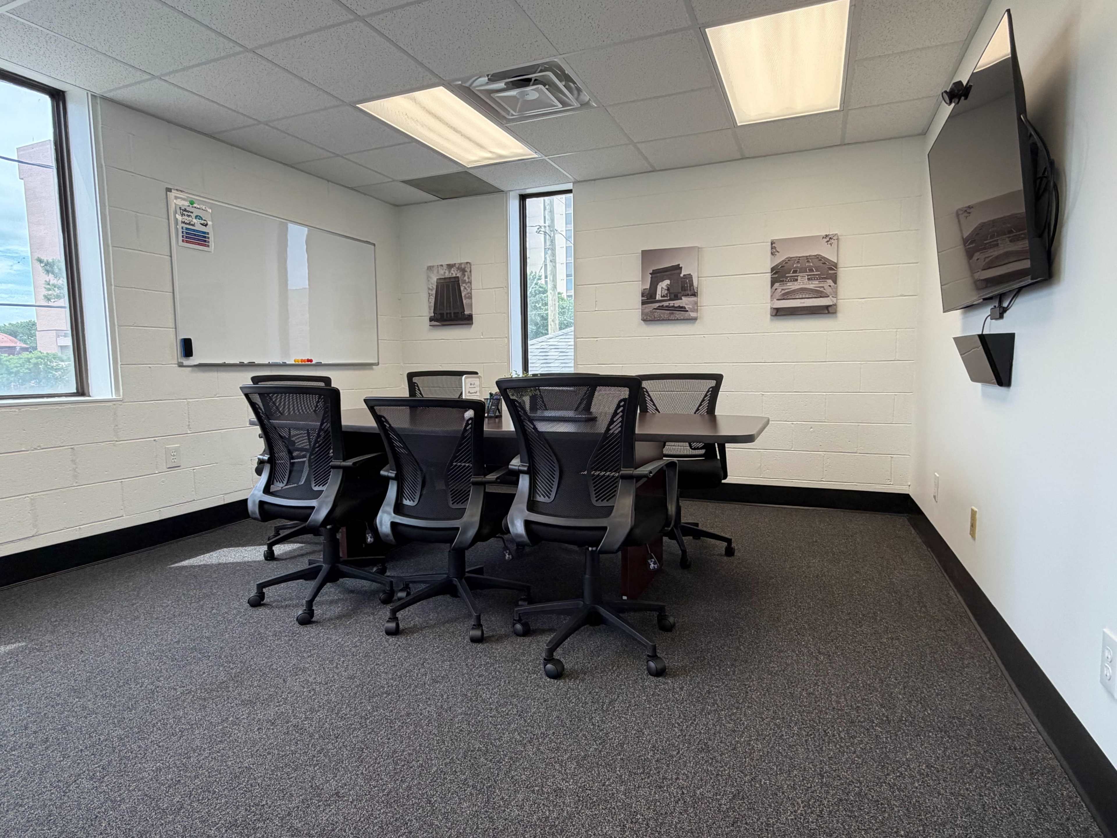 The image shows a conference room with a rectangular table surrounded by six black office chairs, and a wall-mounted TV on one side.