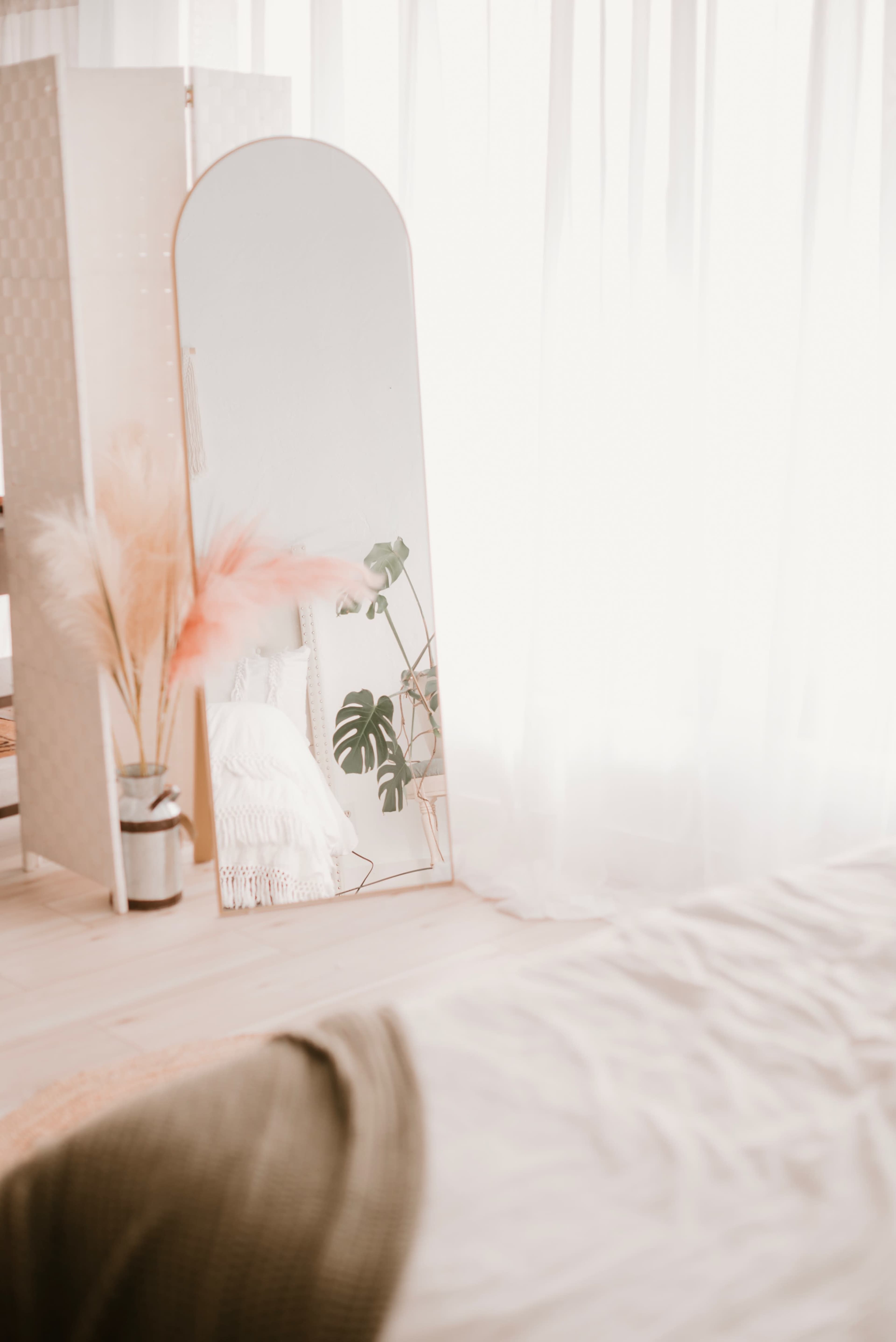 A minimalist bedroom featuring a large mirror reflecting a potted plant and a light fabric partition against a backdrop of sheer curtains.