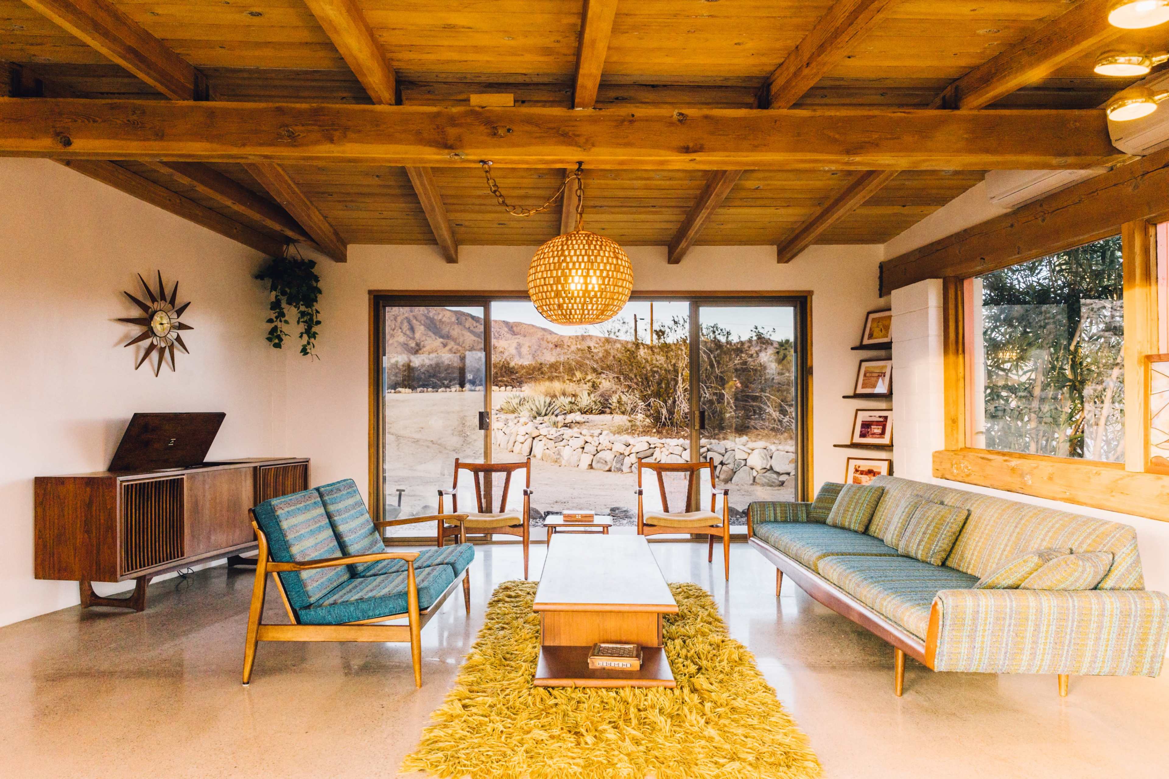 A spacious living room features a wooden ceiling, large windows, a green area rug, and mid-century modern furniture arranged around a coffee table.