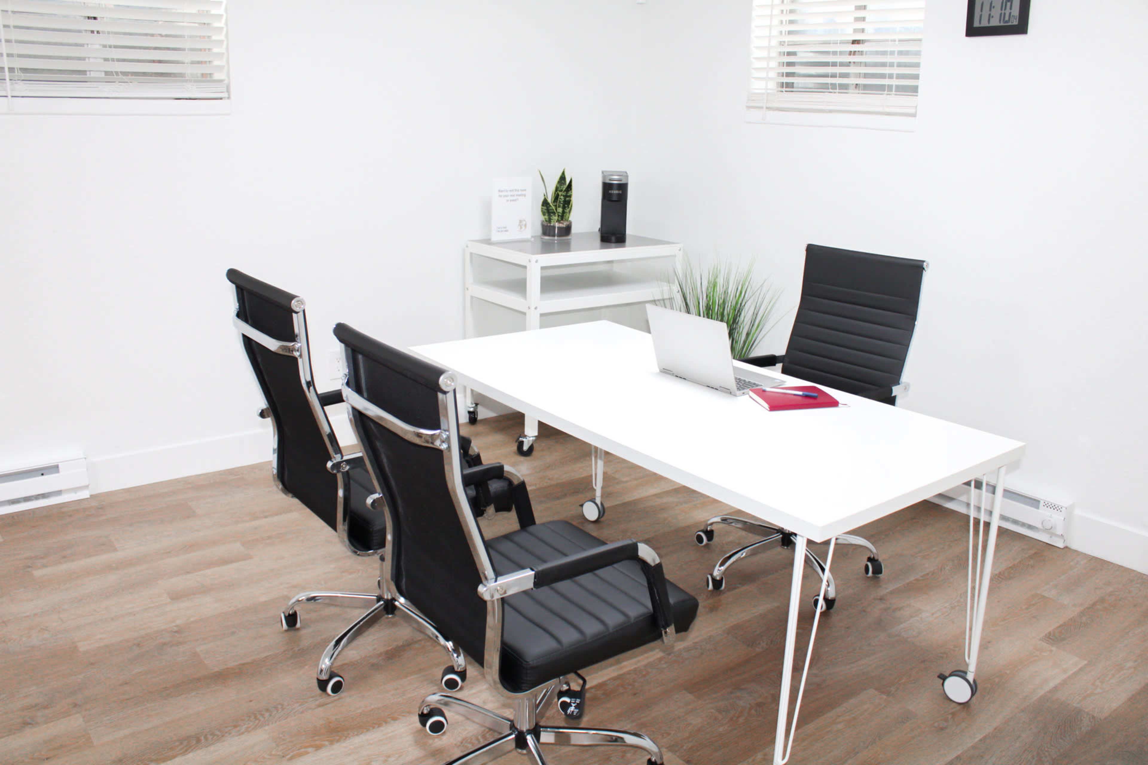 A modern office space features a white table, two black ergonomic chairs, a laptop, and potted plants against white walls.