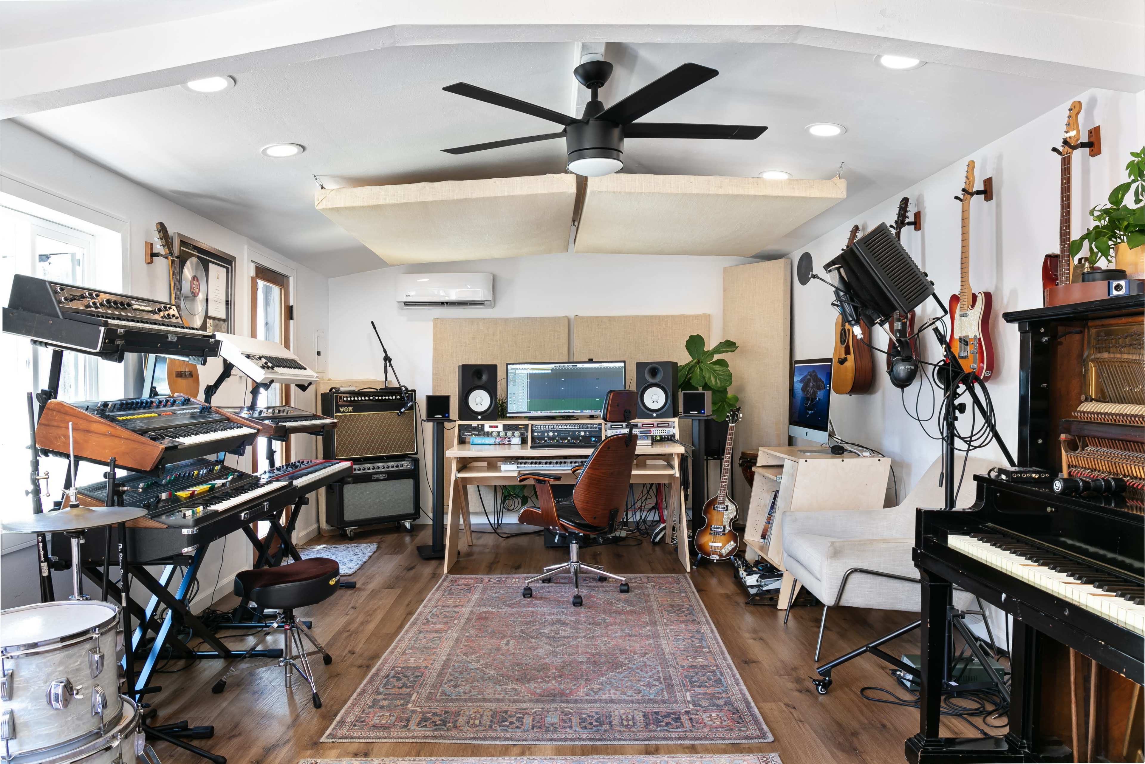 A home music studio featuring various instruments, a computer setup, and sound equipment arranged in a well-lit room with wood flooring.
