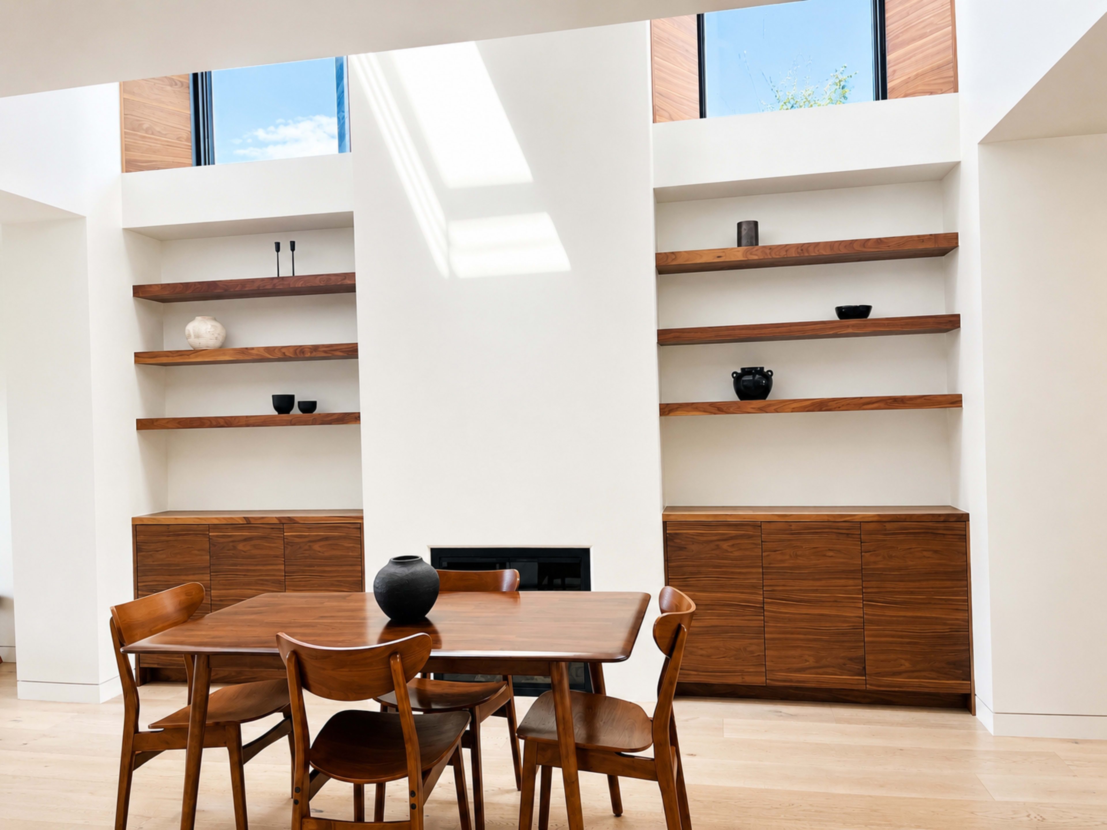 A minimalist dining area features a wooden table and chairs, with open shelving and cabinets against white walls.