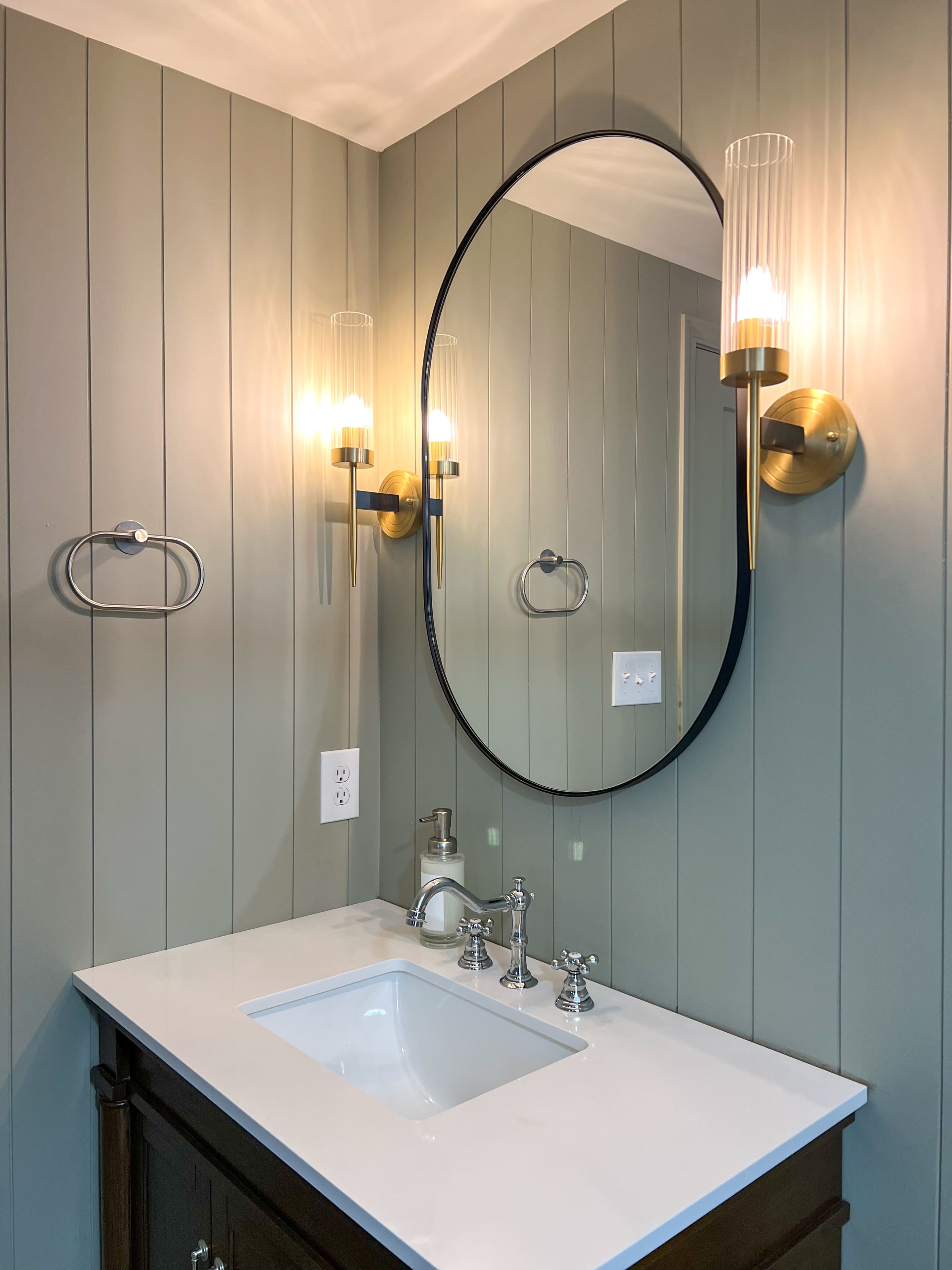 A bathroom with a round mirror above a white sink and a wall-mounted sconces flanking the mirror, all set against green paneled walls.