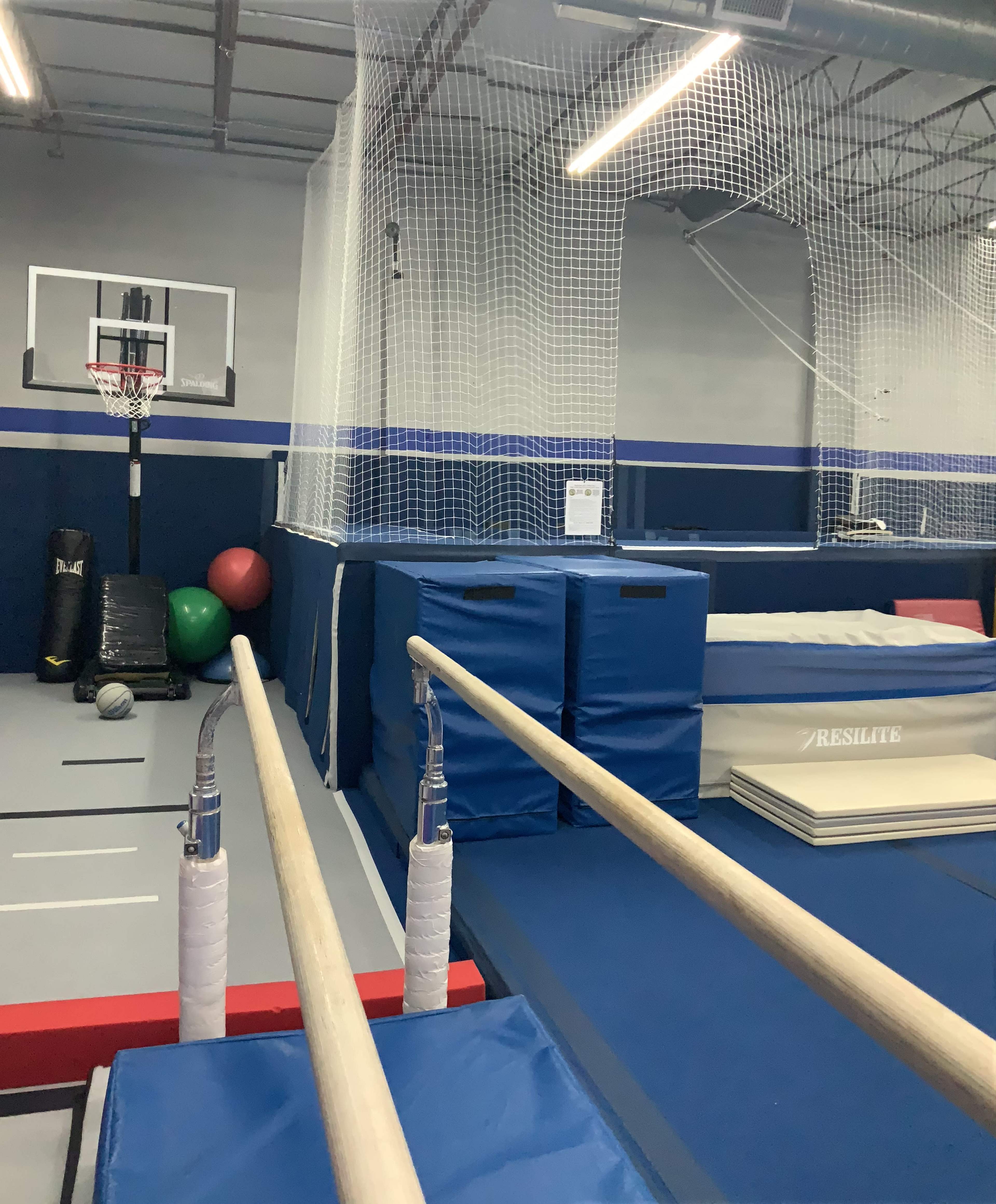 The image shows a gymnastics training facility with parallel bars in the foreground, gym mats, and a basketball hoop in the background.