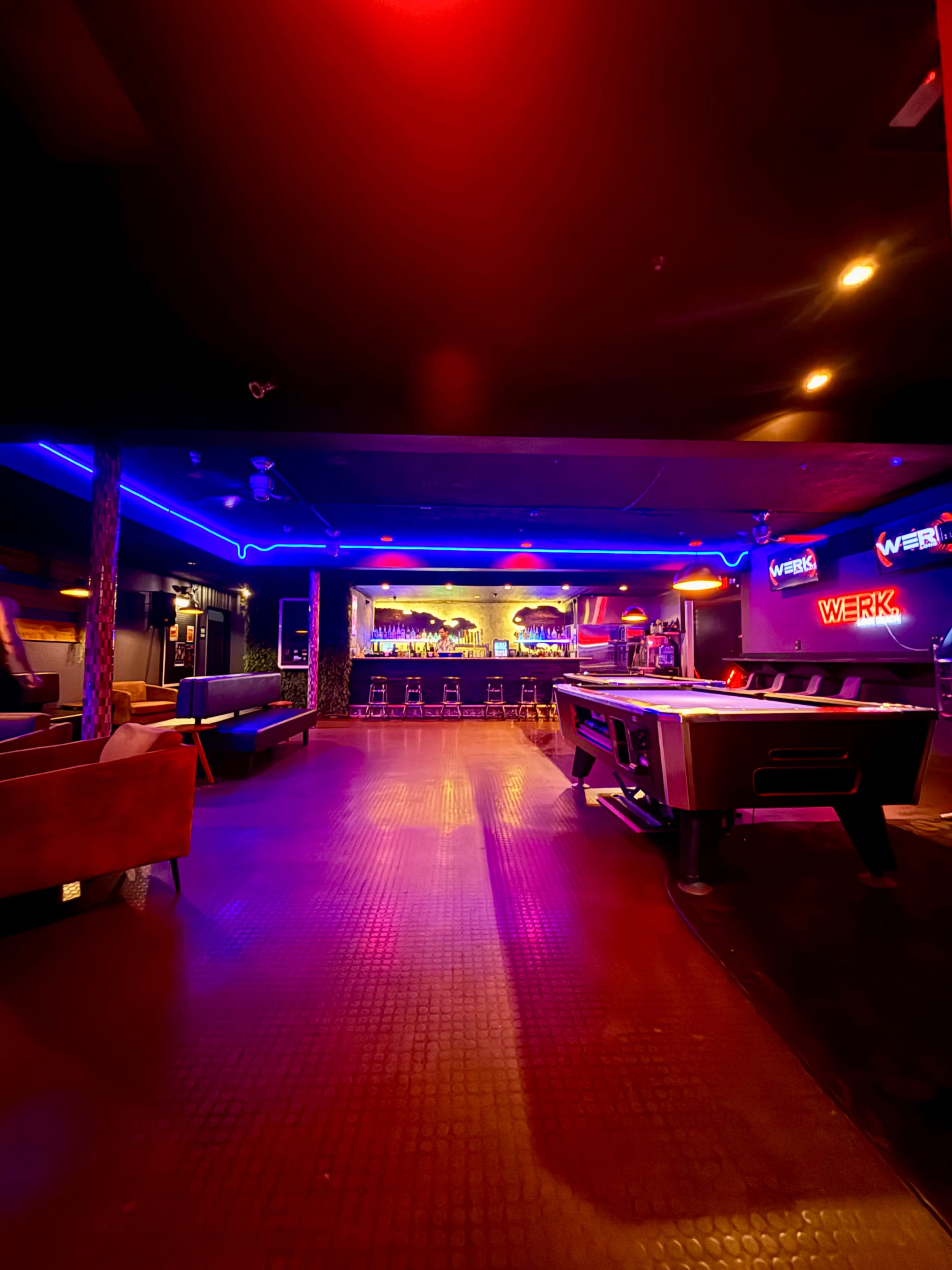 The image shows a dimly lit bar with neon lights, a long bar counter, and a pool table in the foreground.