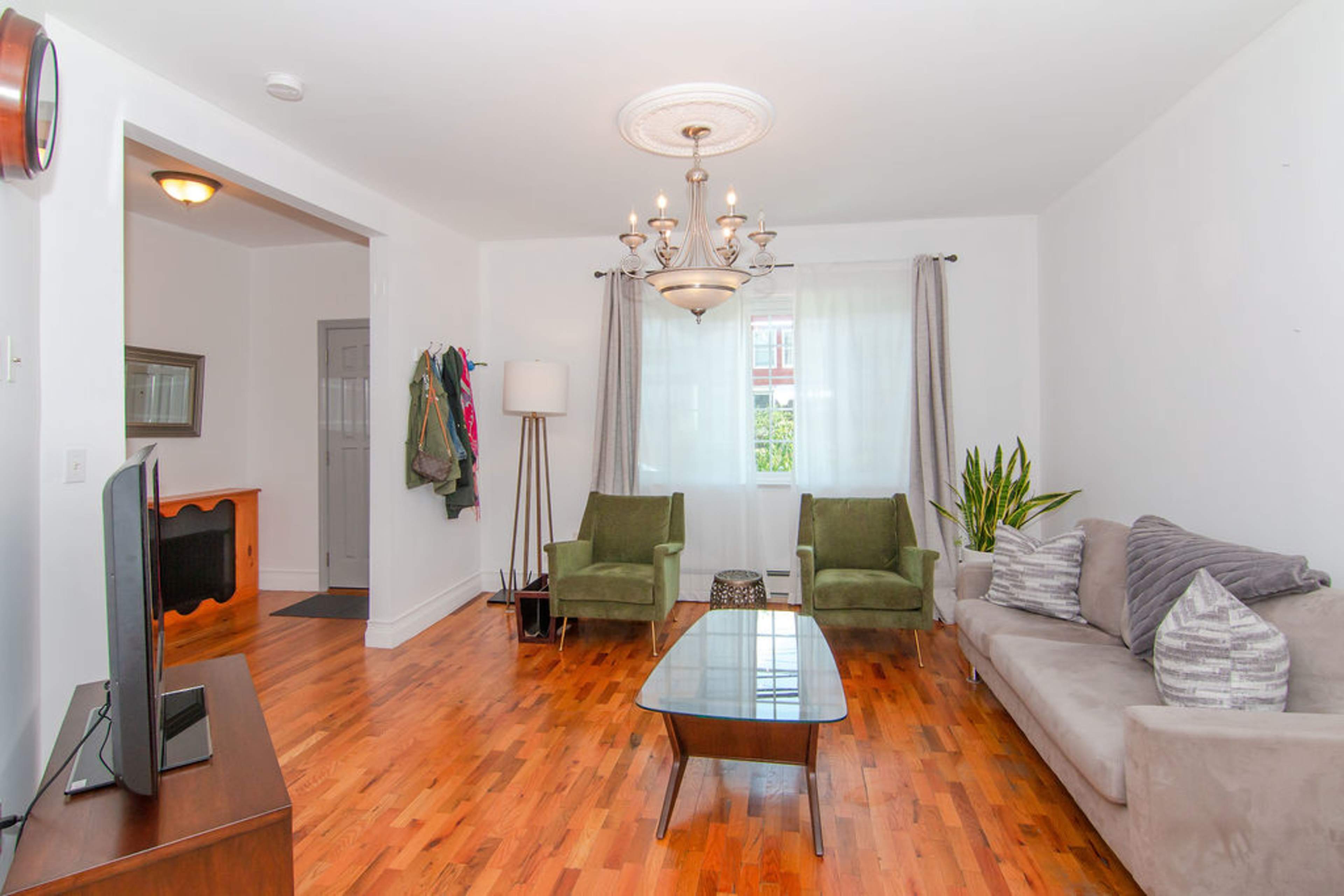 The image shows a living room with two green armchairs, a gray sofa, a coffee table, and a chandelier, illuminated by natural light through the windows.