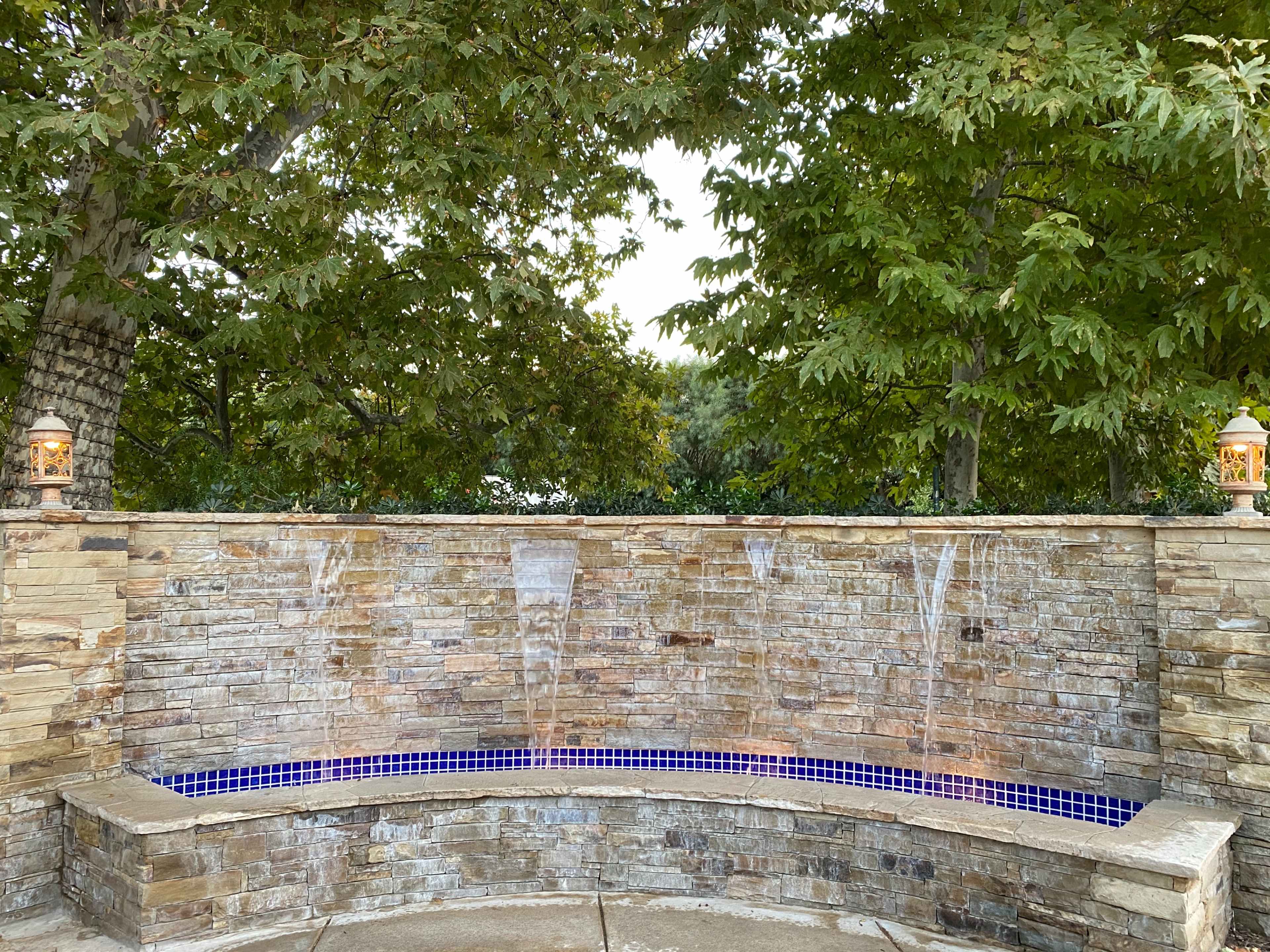 A stone water feature with three streams of water flowing over a curved wall, surrounded by greenery and lanterns.