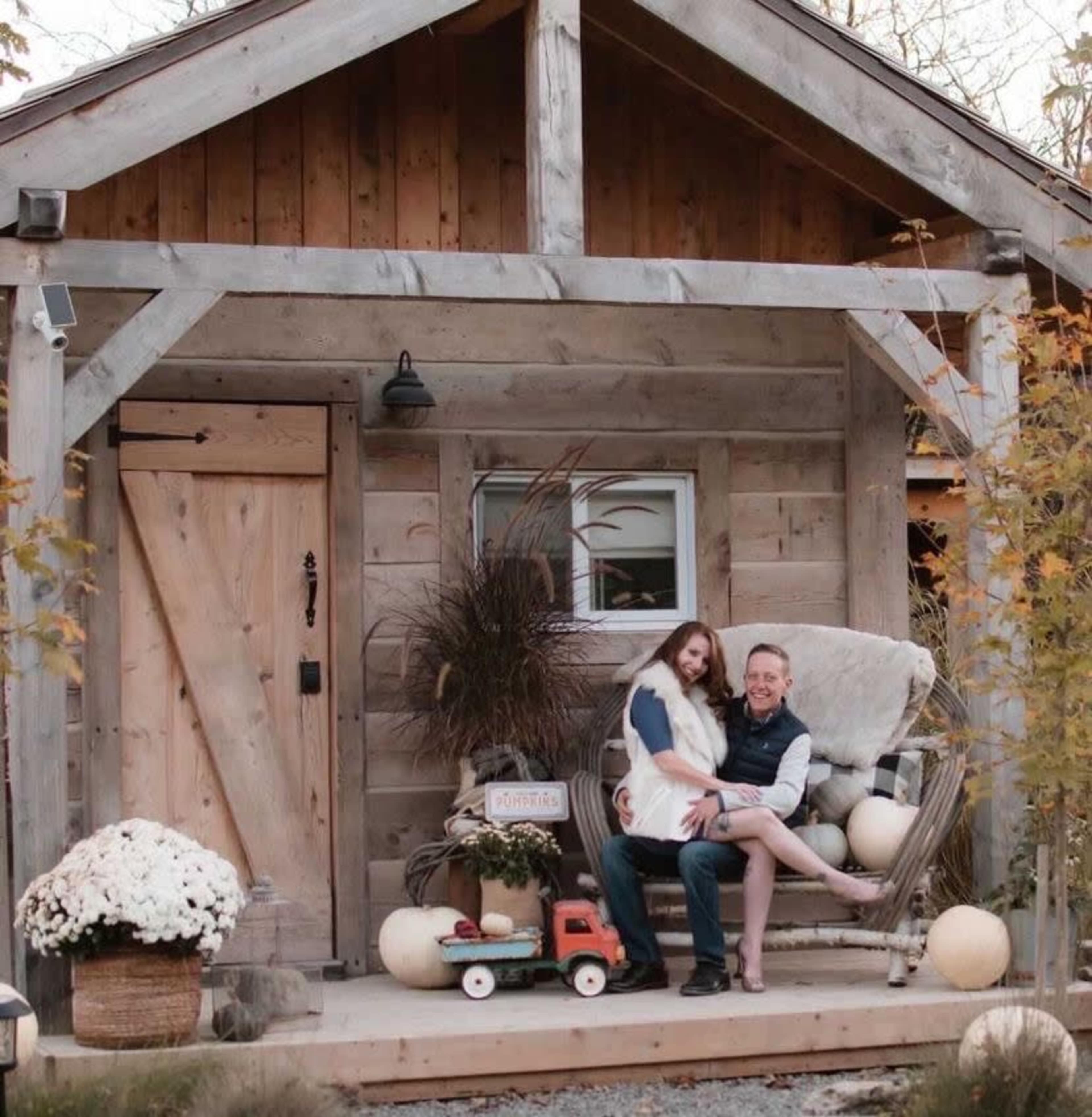 A couple sits on a rocking chair on the porch of a rustic wooden cabin, surrounded by decorative pumpkins and flowers.