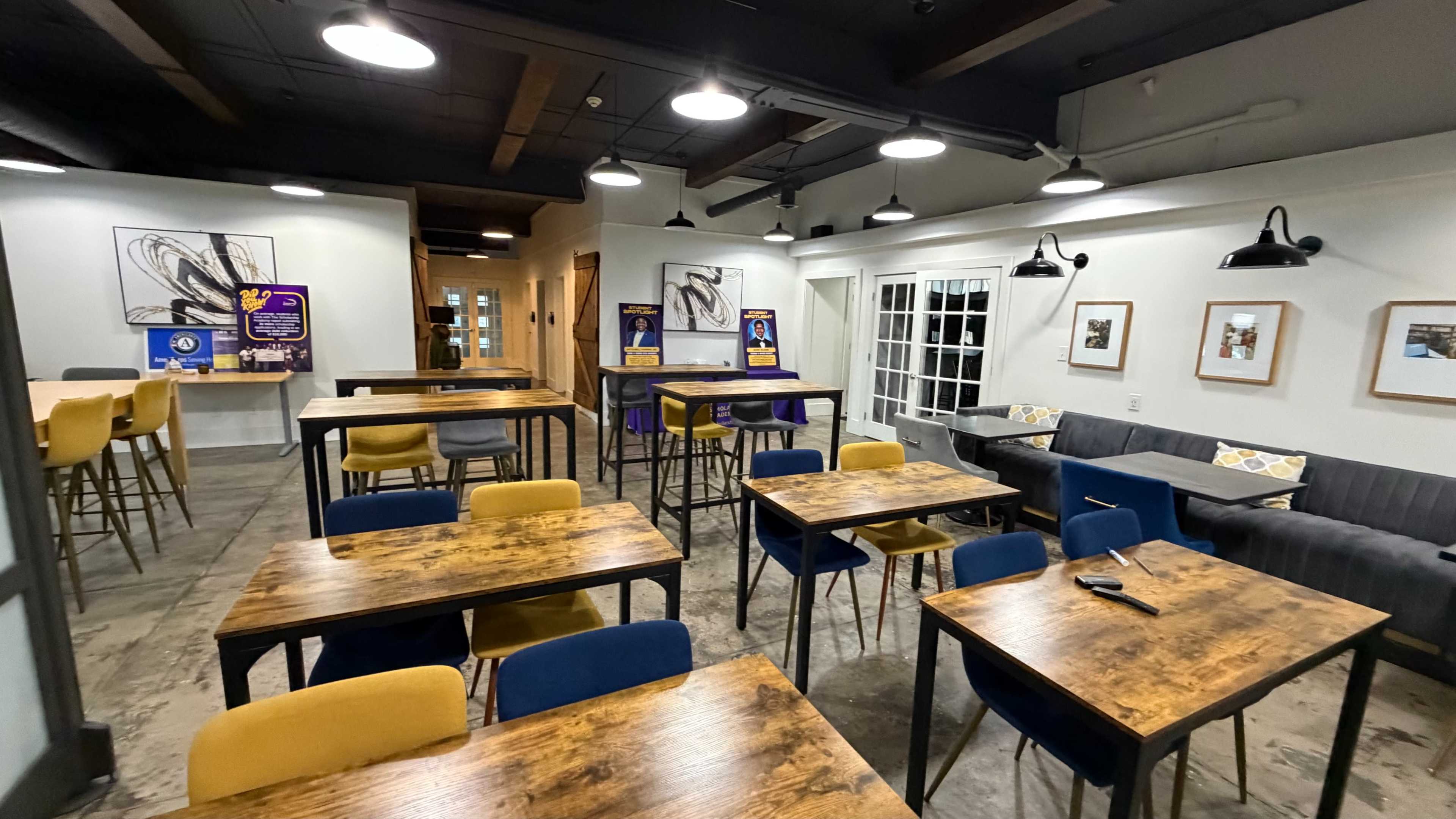 The image shows a modern café interior featuring wooden tables, colorful chairs, and artwork on the walls, with a cozy seating area in the background.