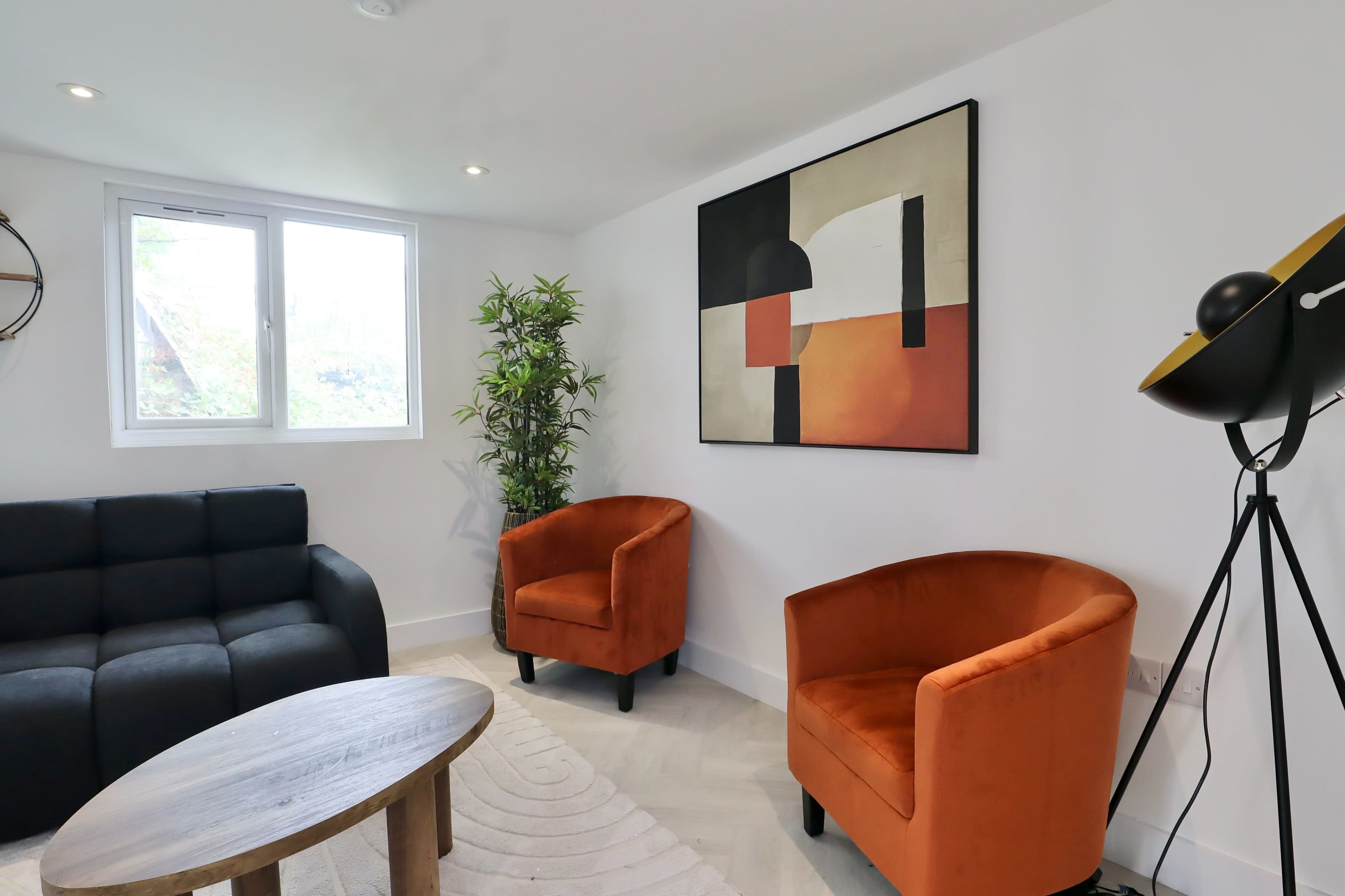 The image shows a modern living room with a black sectional sofa, two orange armchairs, a wooden coffee table, a decorative plant, and abstract wall art.