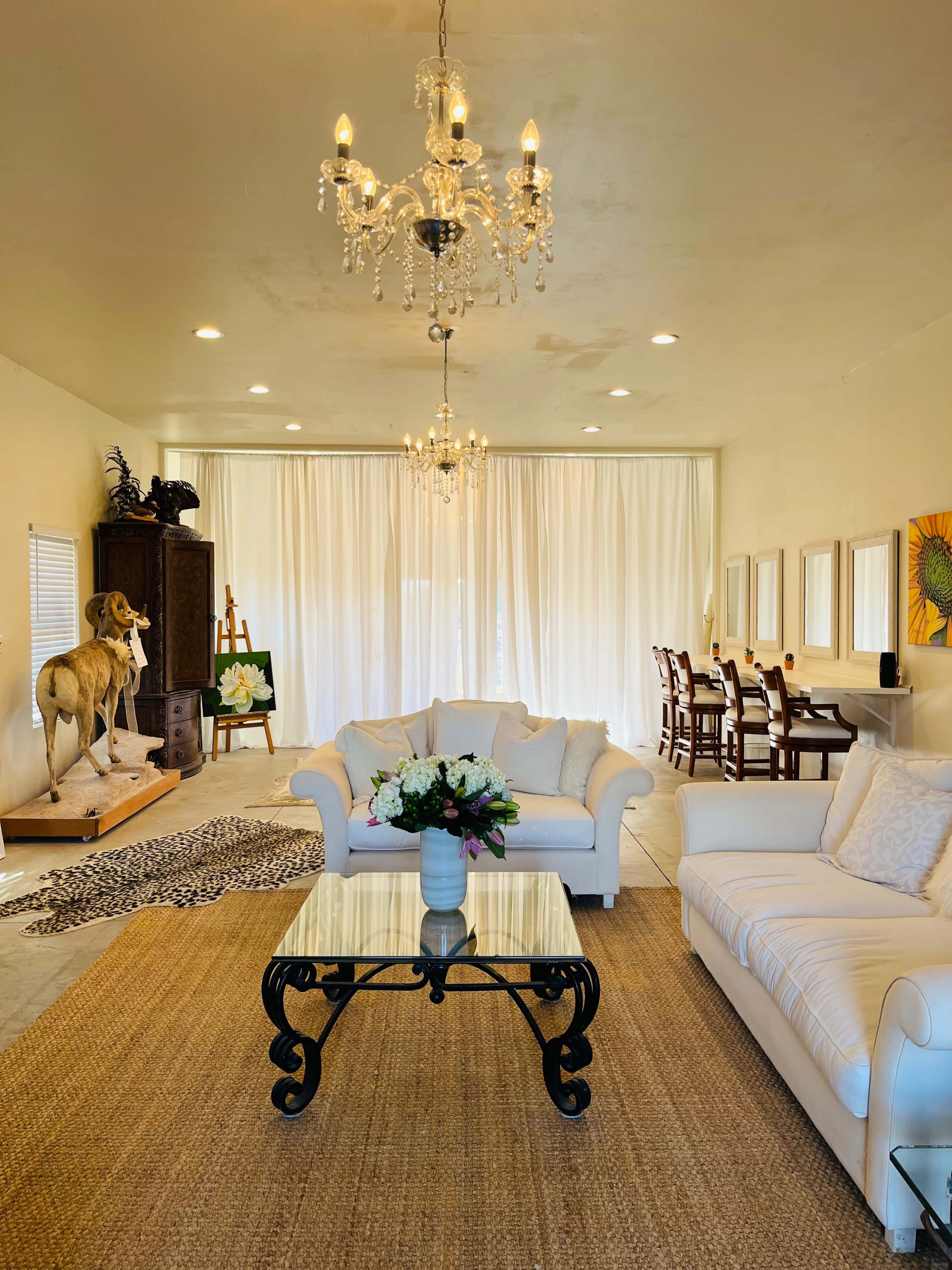 A bright living space with a white sofa, a glass coffee table, a decorative rug, and a dining area featuring a wooden table and chairs under chandeliers.