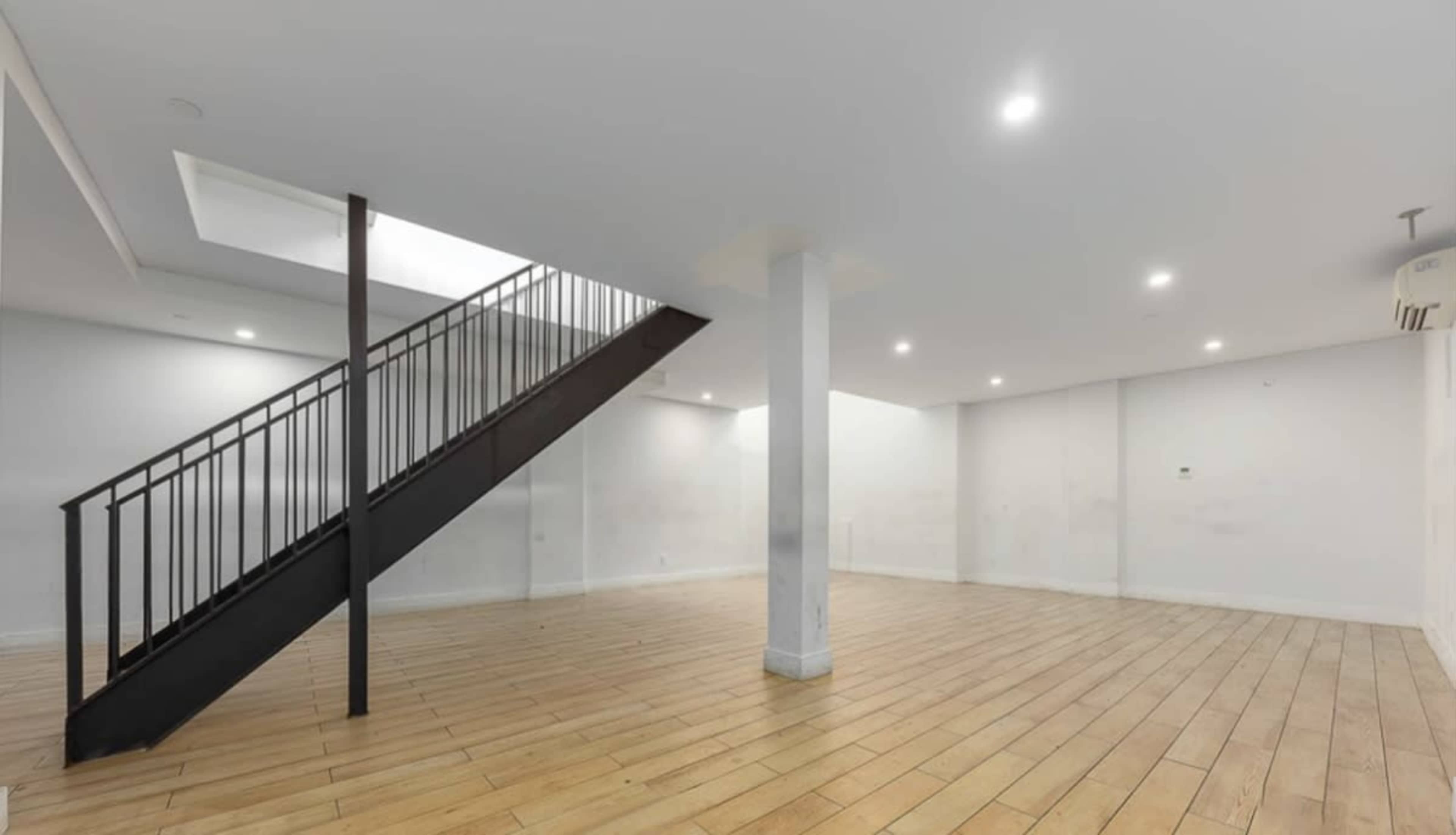 An empty room with wooden flooring and a staircase on the left leading to an upper floor, illuminated by recessed lighting.