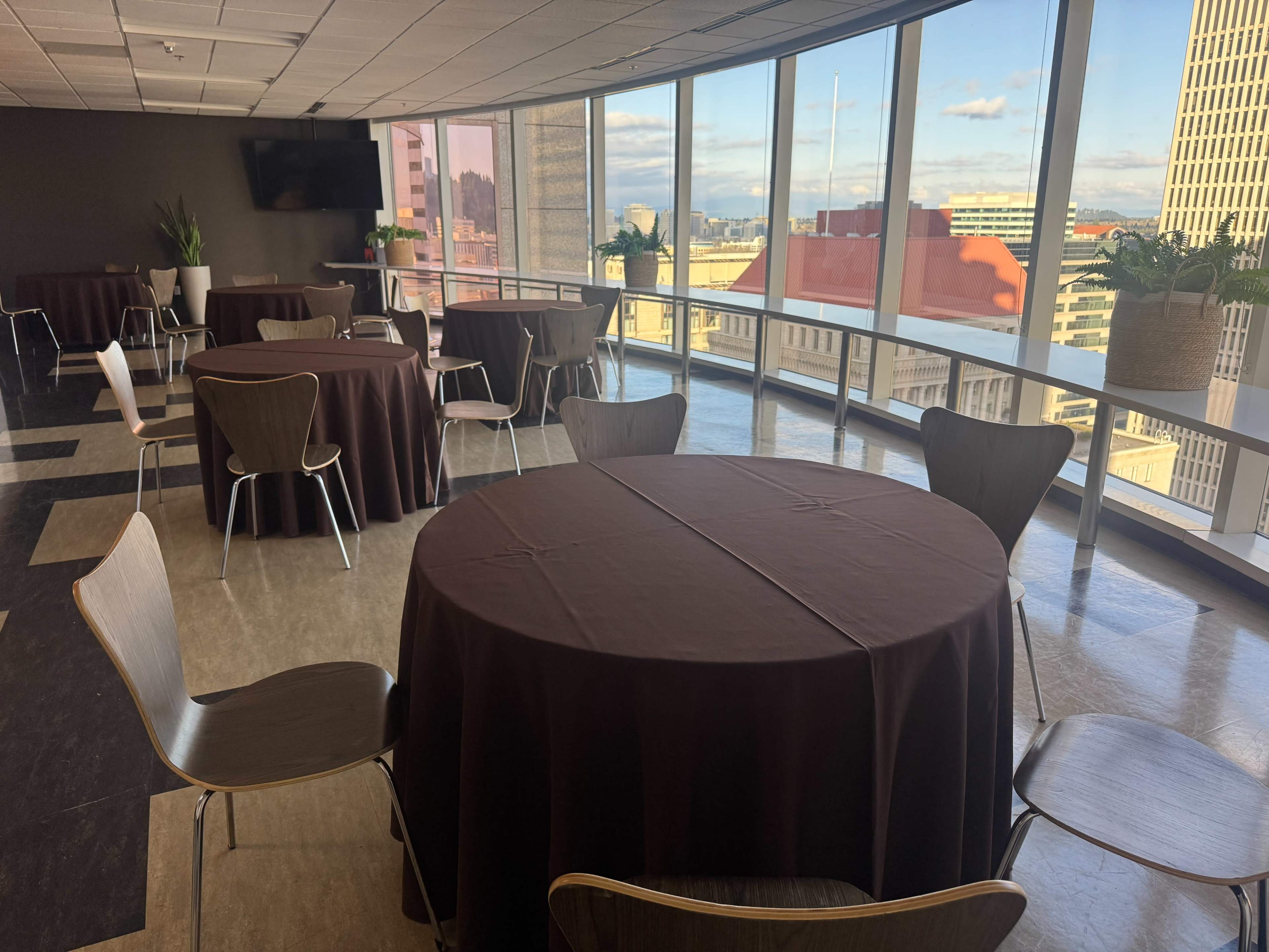 A spacious indoor venue features round tables with dark tablecloths and modern chairs, overlooking a cityscape through large windows.