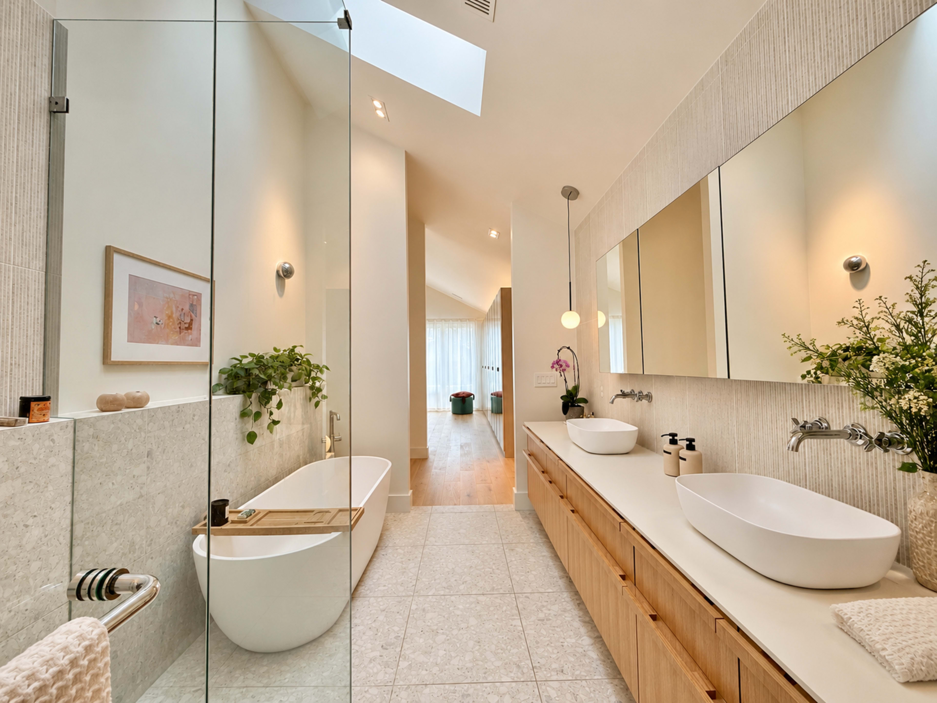 The image shows a modern bathroom featuring a freestanding bathtub, dual sinks on a long wooden vanity, and a large mirror, with natural light streaming in from a skylight.