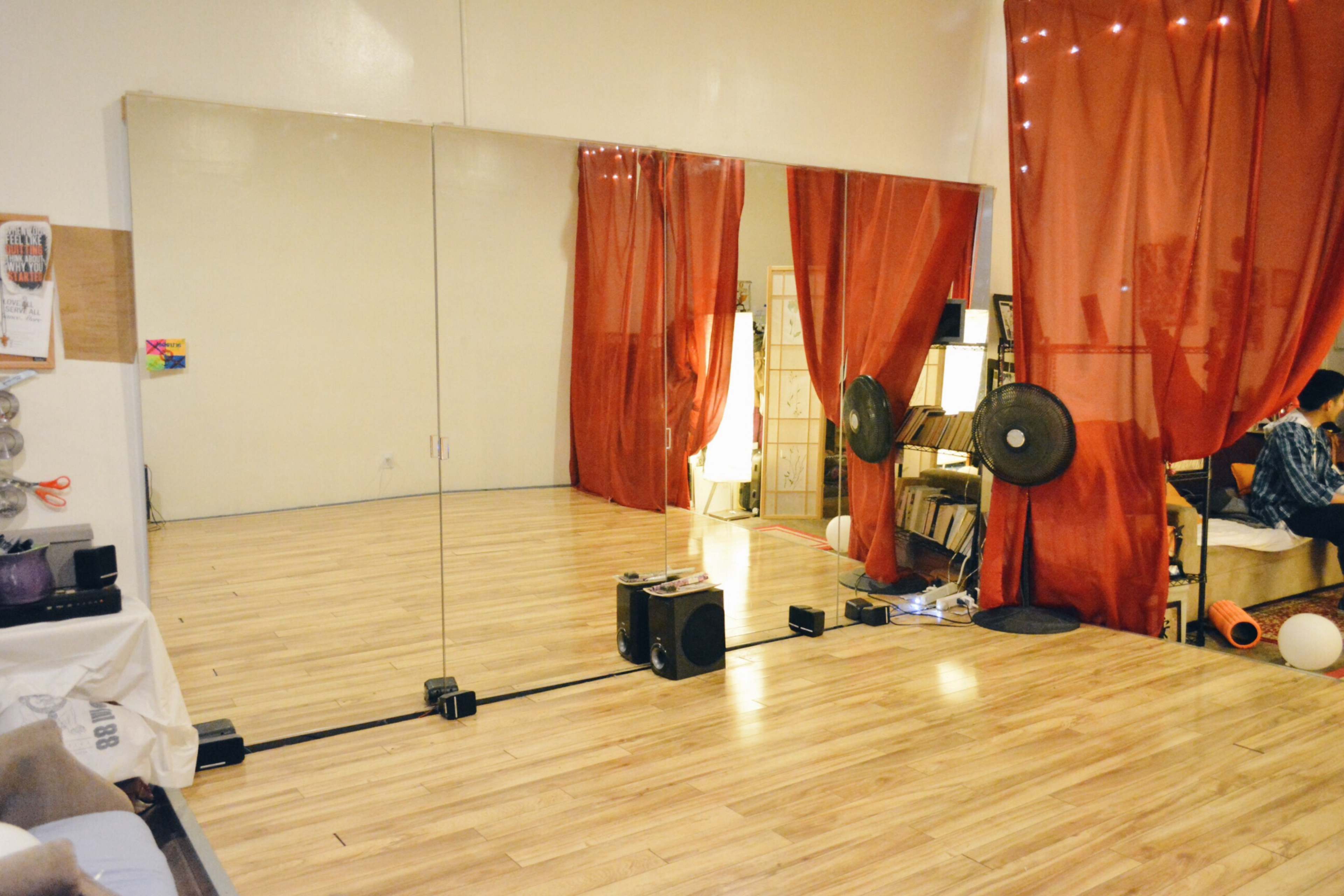 A spacious dance studio with wooden flooring, a large mirror on one wall, and red curtains, along with shelves and fans in the corner.