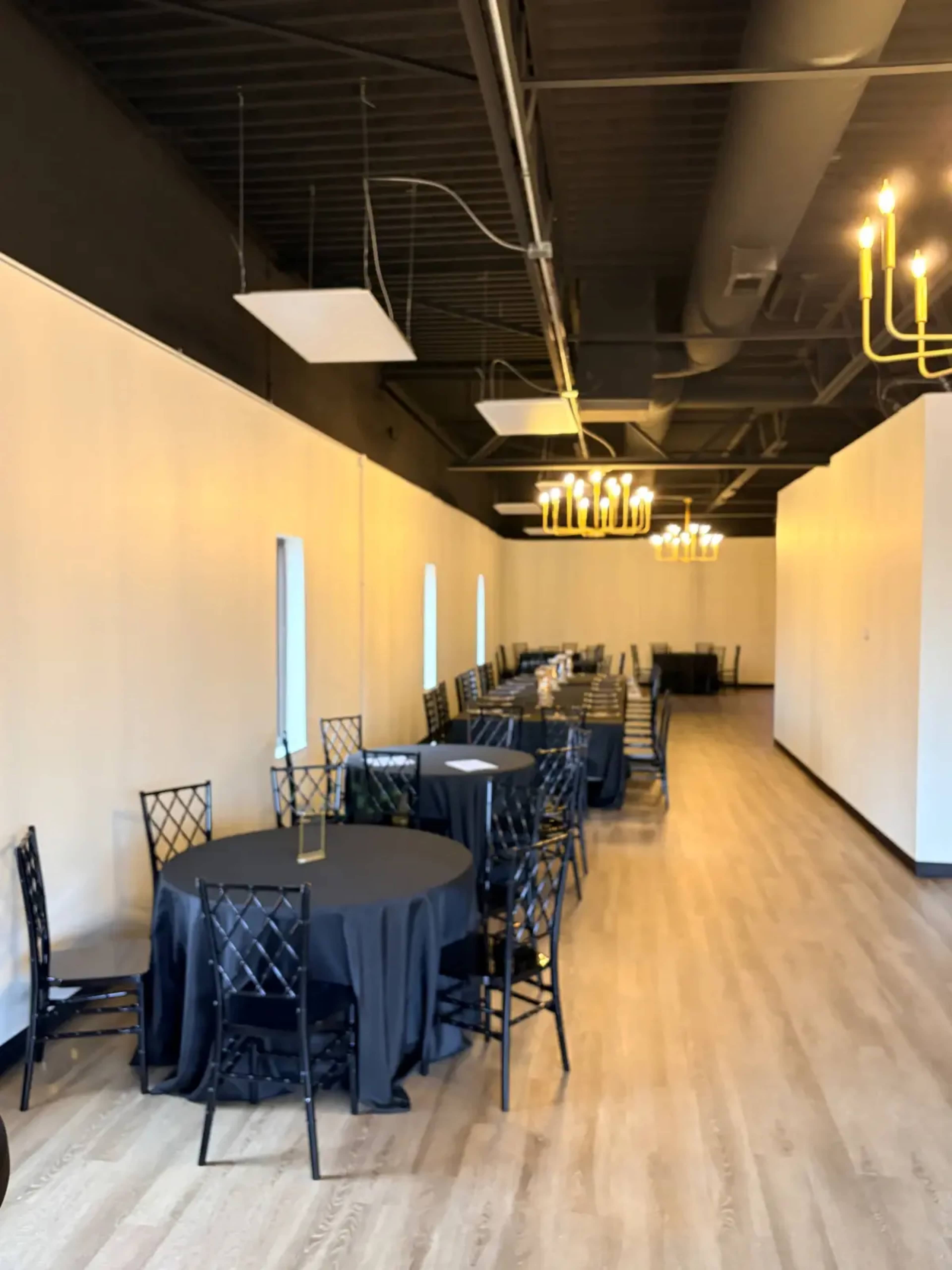 The image shows a long banquet hall with black tablecloth-covered tables arranged in rows, illuminated by modern chandeliers.