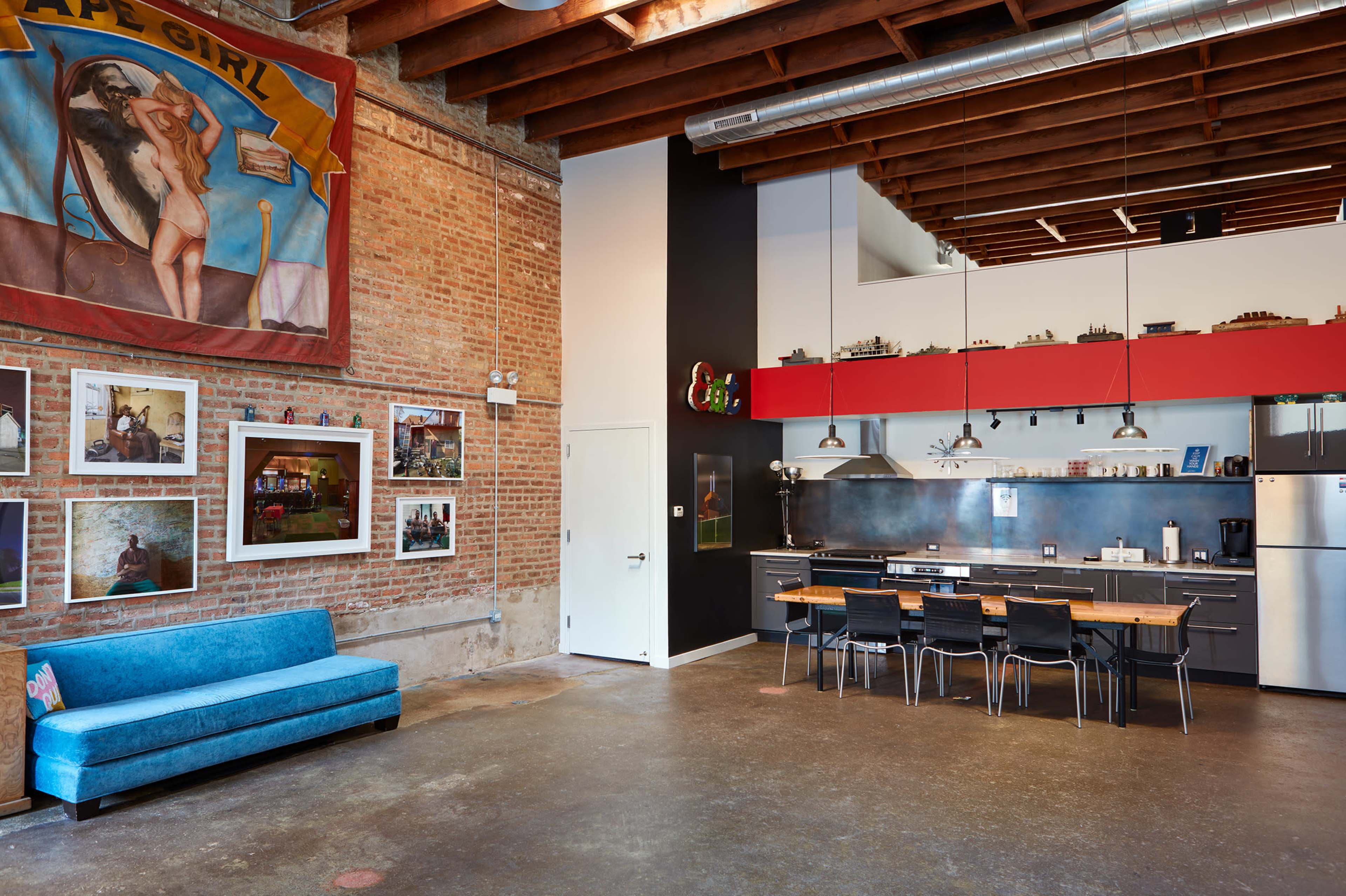 A spacious industrial-style kitchen and dining area with a brick wall, colorful artwork, and a long table surrounded by black chairs.