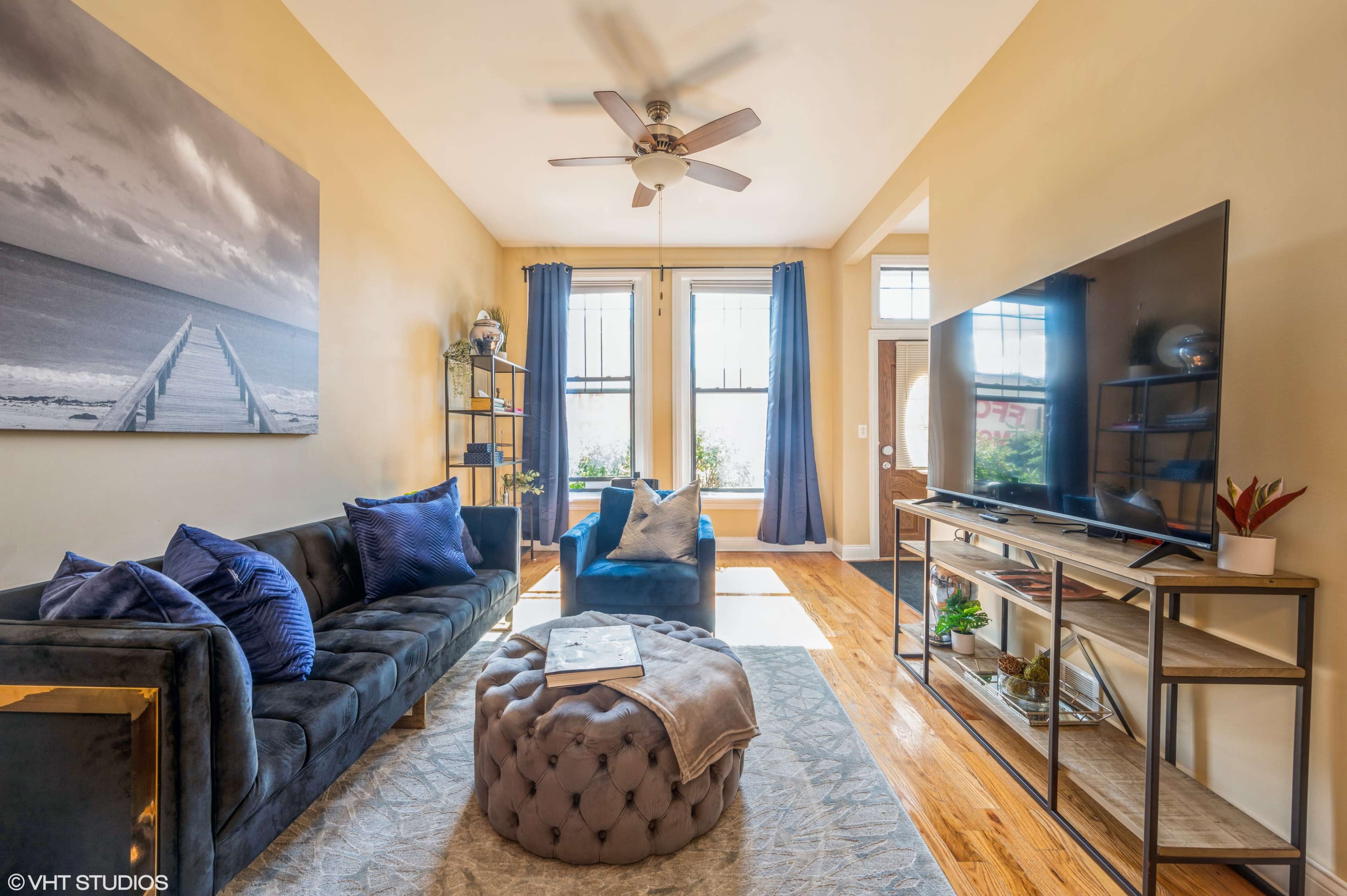 A brightly lit living room with a blue sofa, a decorative ottoman, and a large television on a stand against a wall.