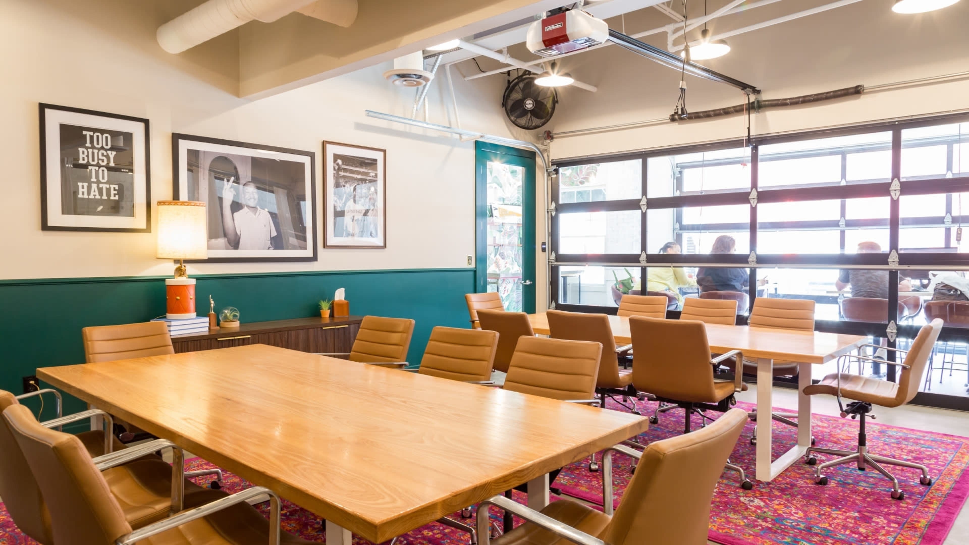 A conference room features a large wooden table surrounded by various chairs, with framed artwork on the walls and a large garage door opening to a view of people outside.