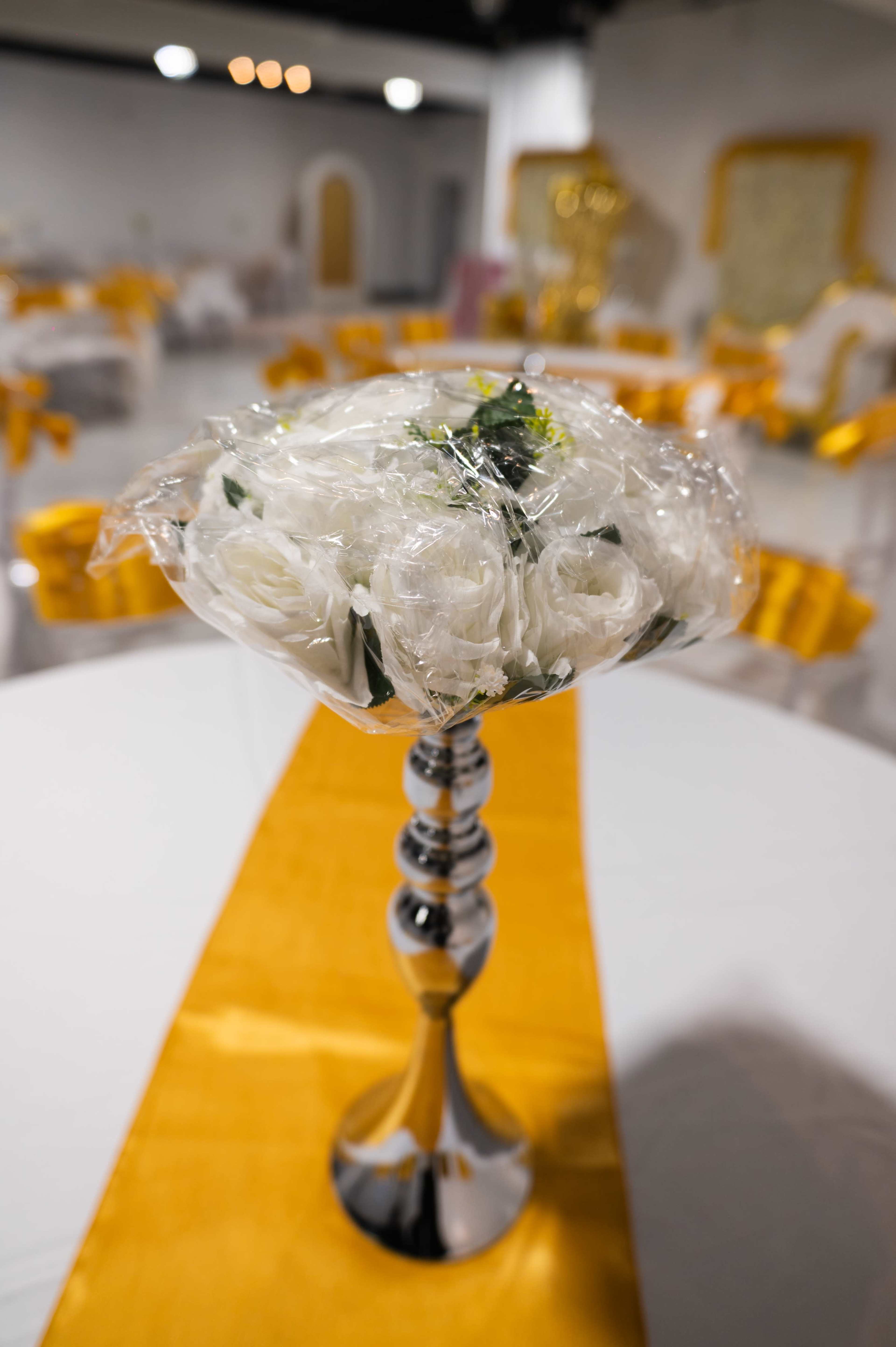 A bouquet of white flowers is displayed in a silver vase on a table with a yellow table runner in a decorated event space.