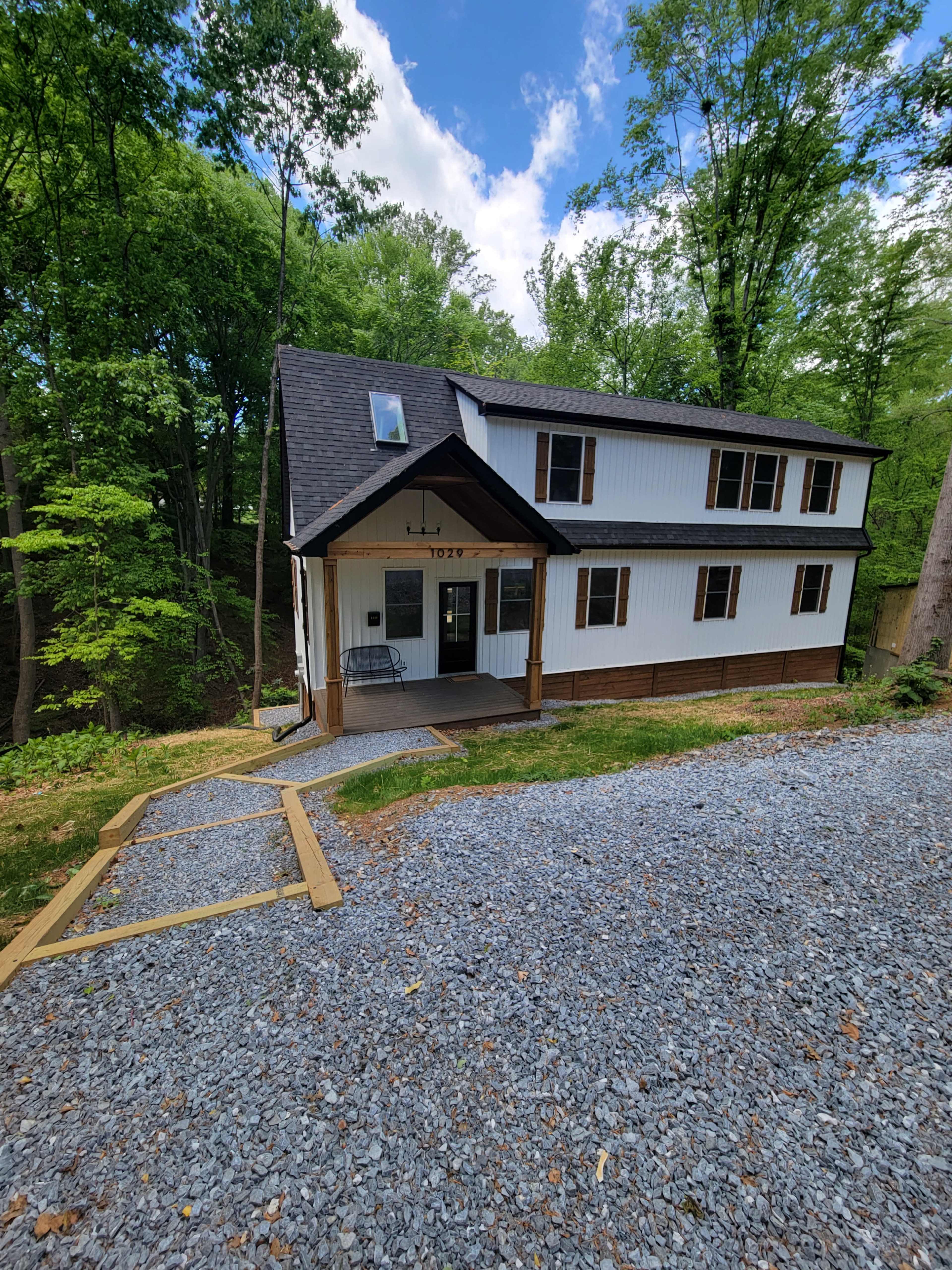 A two-story house with white and brown siding is situated among green trees, with a gravel path leading to the front porch.