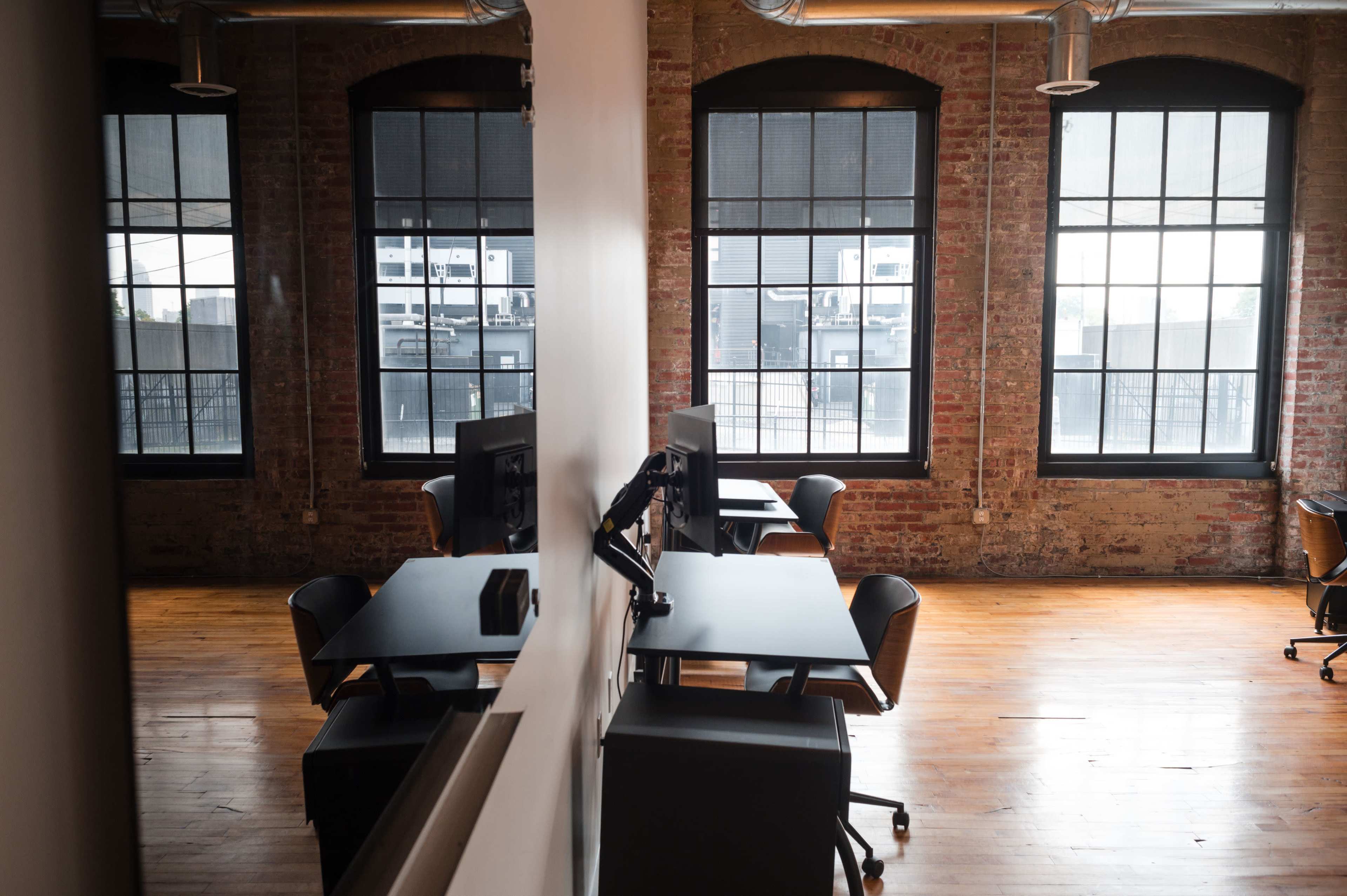 A modern office space with wooden floors, exposed brick walls, and several desks facing large windows.