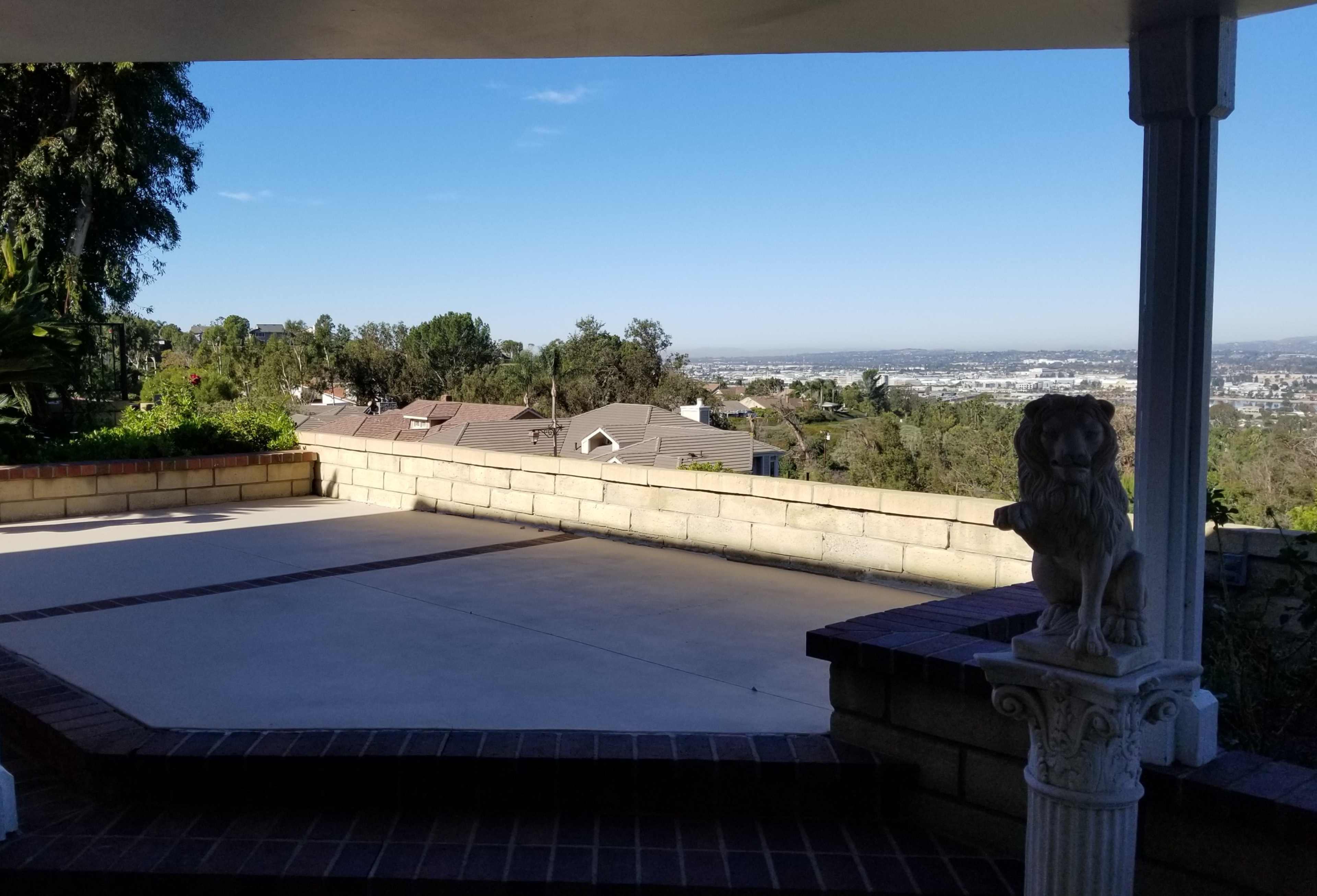 The image shows a stone patio with a lion statue in the foreground, overlooking a view of houses and a valley under a clear blue sky.