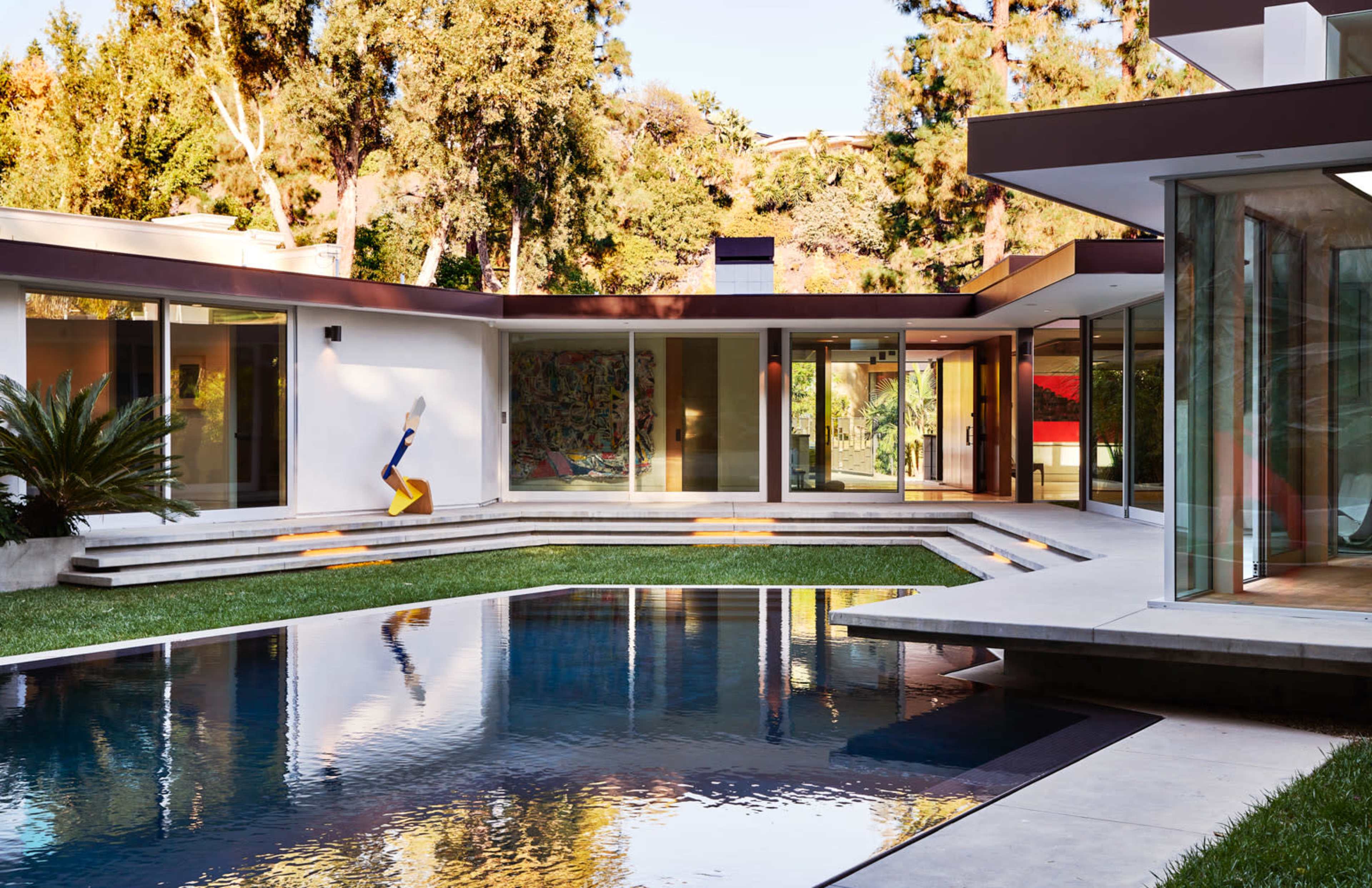 A modern house with large glass walls surrounding a rectangular swimming pool and landscaped yard.