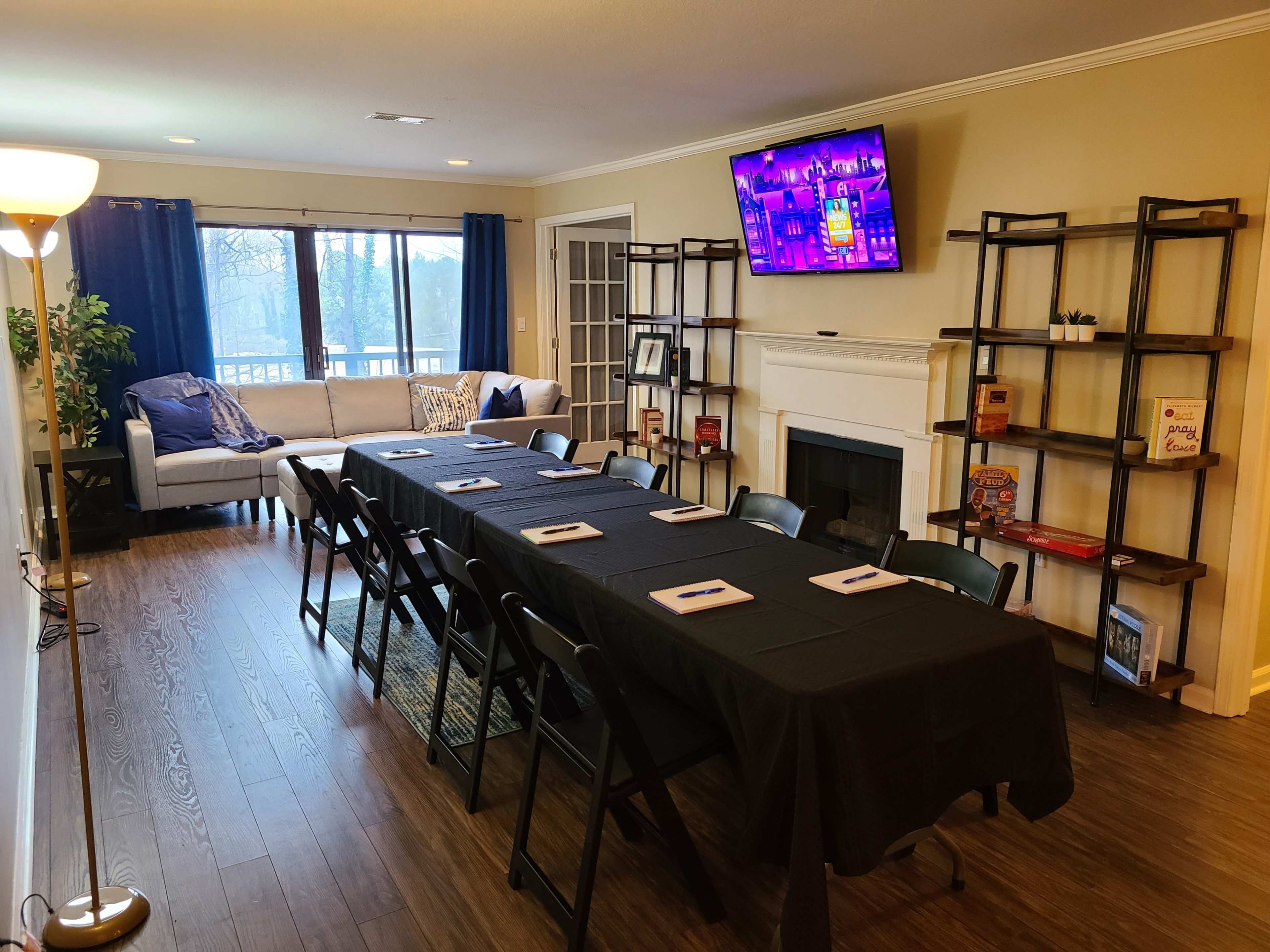 A meeting room features a long black table set with notepads, chairs, a television mounted on the wall, and a sofa in the background.