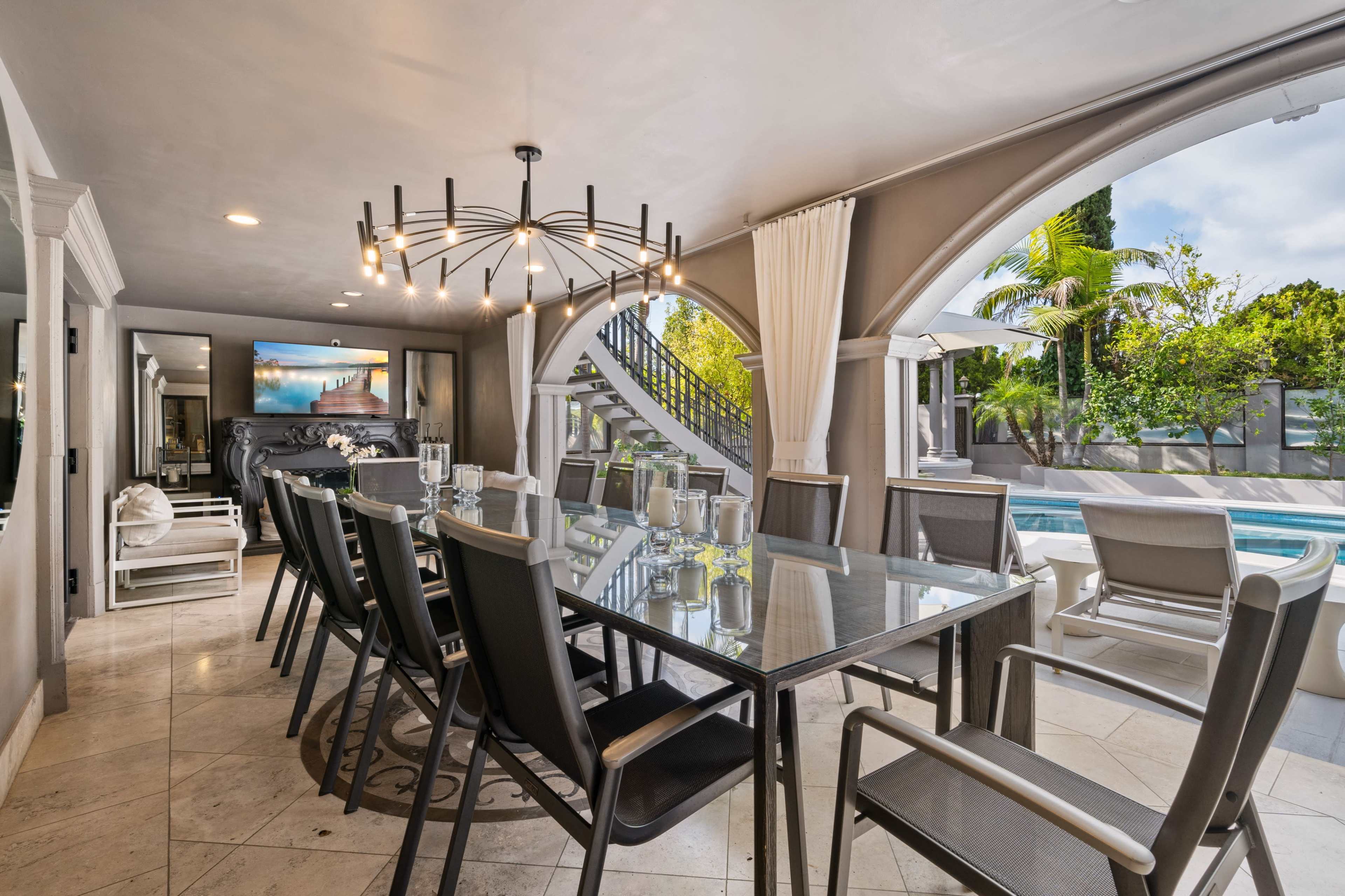 The image shows a dining area with a long glass table surrounded by chairs, large windows, and a view of a pool and outdoor seating area.